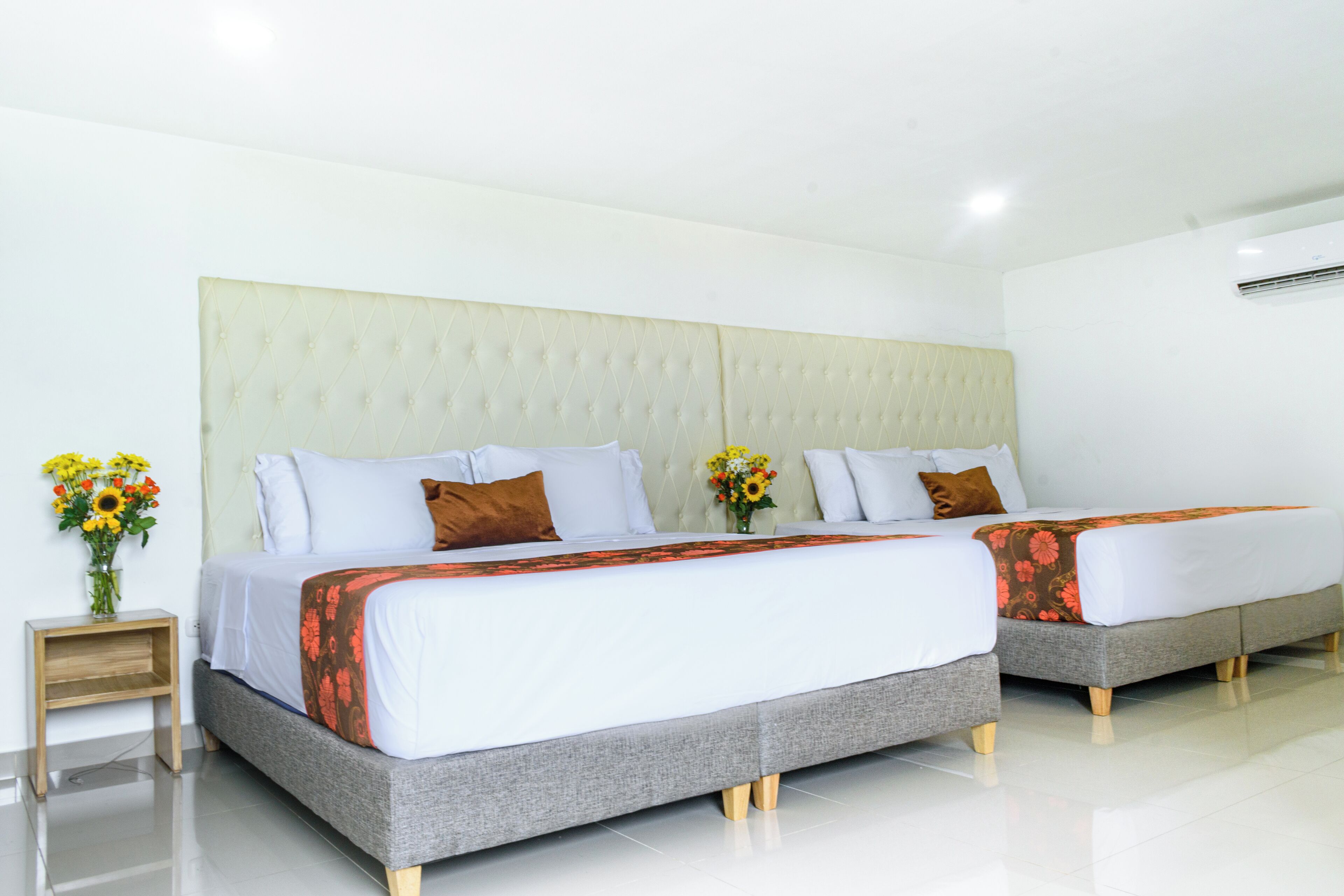 Habitación de hotel con dos camas, un cabecero grande y acolchado, y una mesita de noche pequeña con un jarrón de flores.