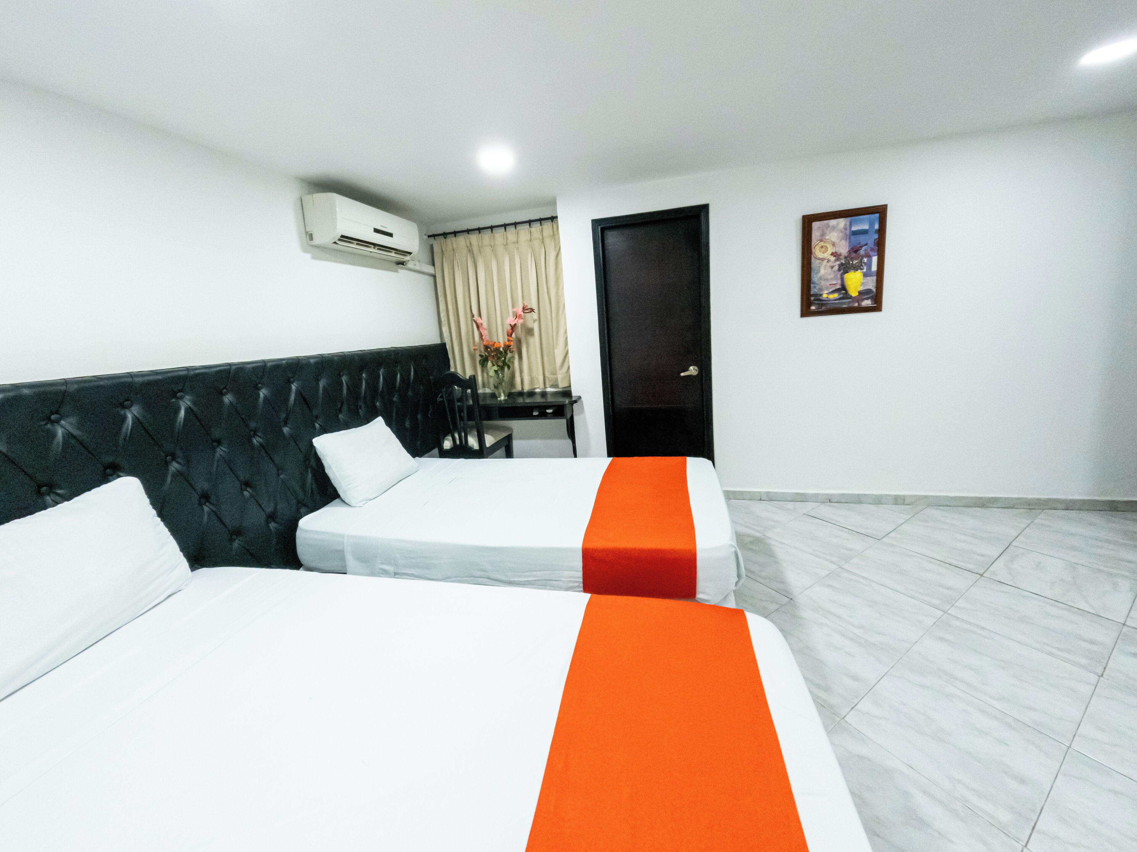 Habitación de hotel con dos camas, un escritorio, una silla, equipo de aire acondicionado, una puerta y un cuadro en la pared.