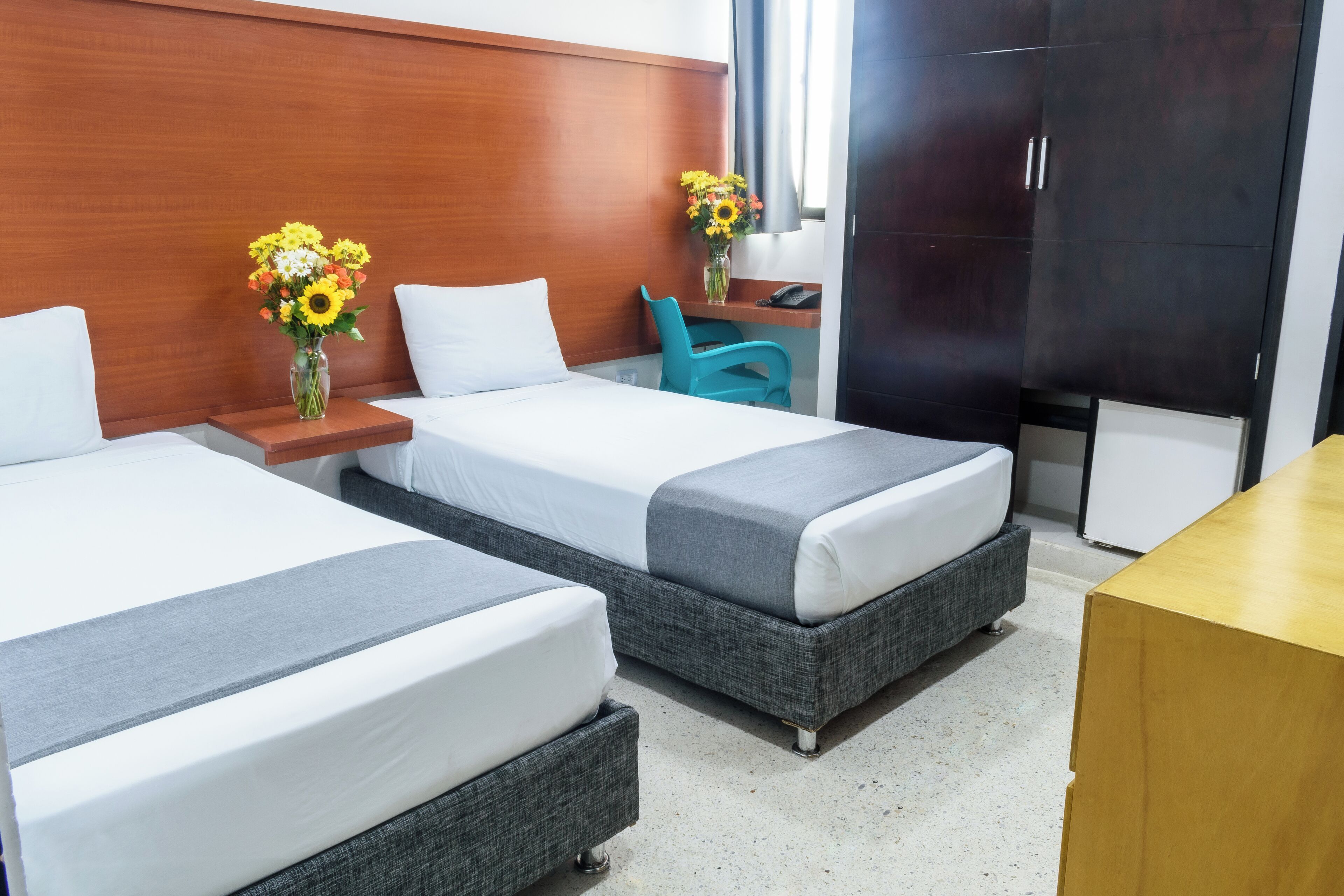 A hotel room with two beds, a wooden headboard, a desk with a chair, a vase with flowers, and a wardrobe.