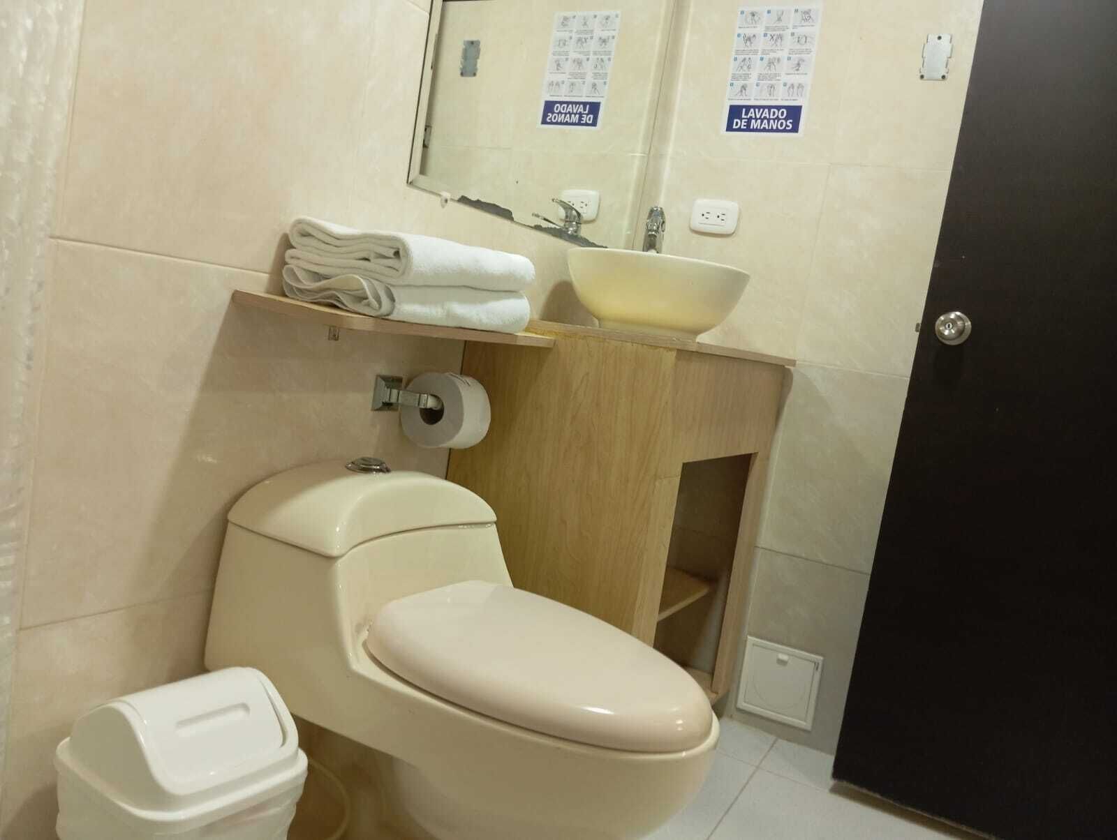 Un baño con inodoro, lavamanos y espejo. Hay toallas sobre el lavamanos y un dispensador de toallas de papel.