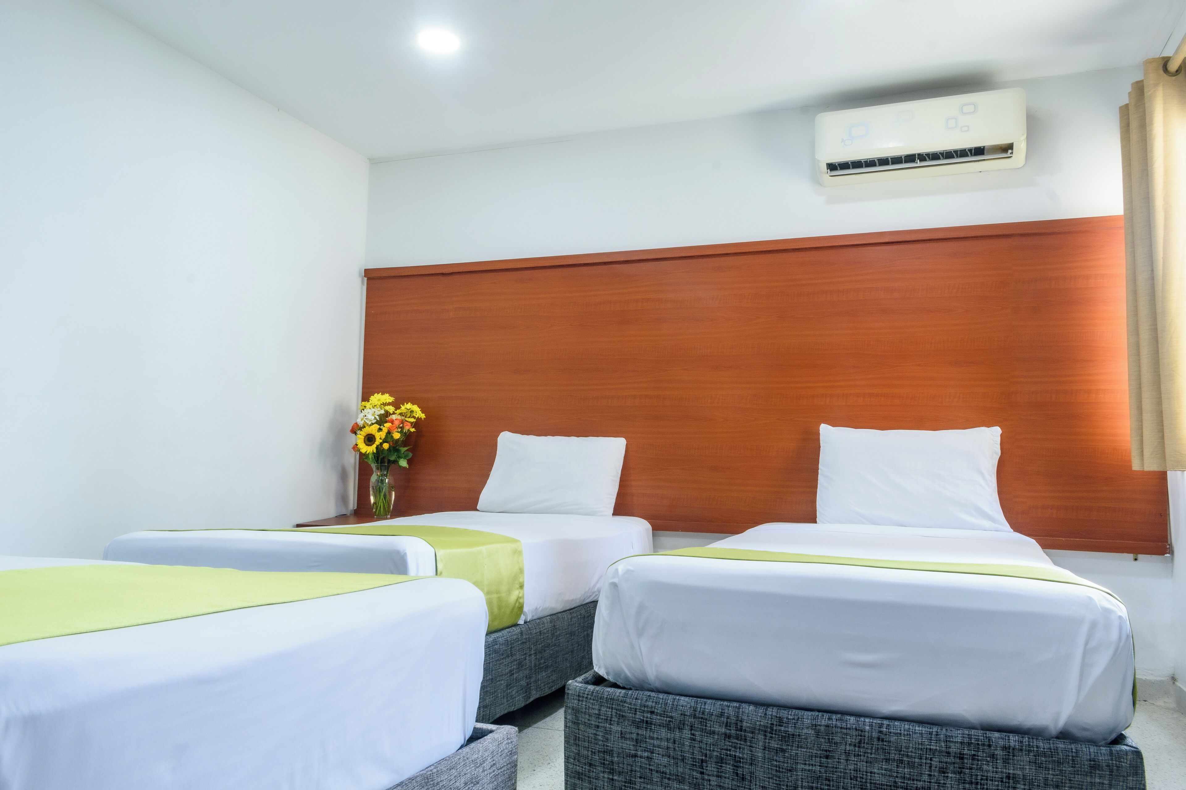 Habitación de hotel con dos camas, cabecera de madera, unidad de aire acondicionado y un jarrón con flores.