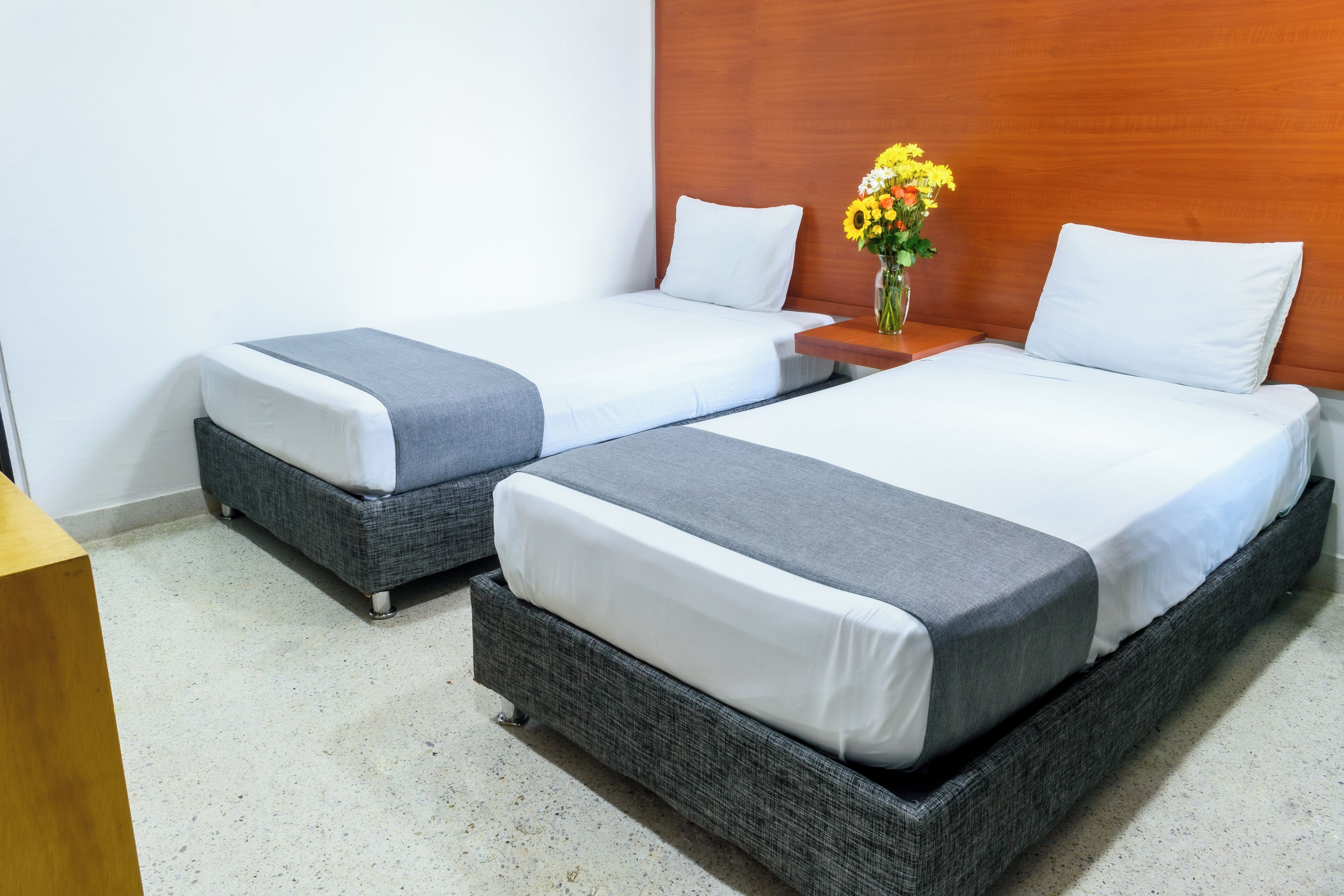 A hotel room with two single beds, a wooden headboard, a small table with a vase of flowers, and a wooden nightstand.