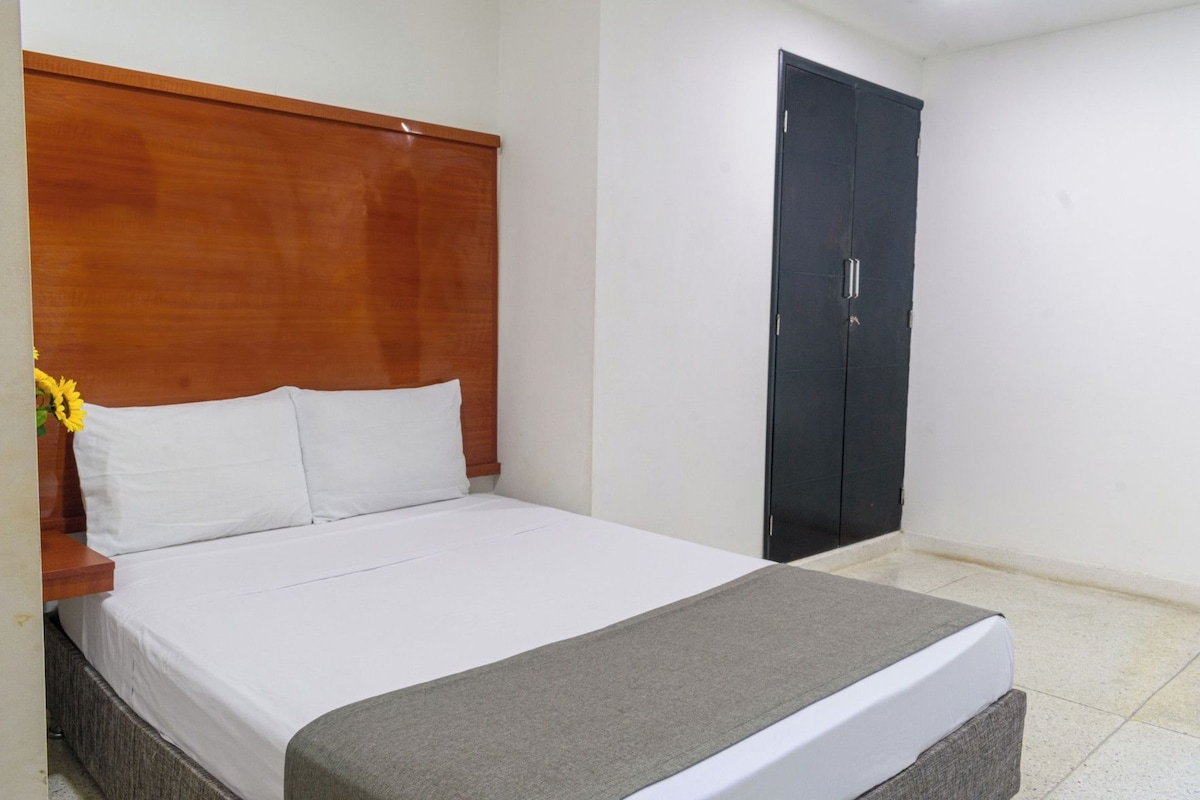 A hotel room with a bed, a wooden headboard, a grey throw, a black wardrobe, and a small table with a vase of sunflowers.