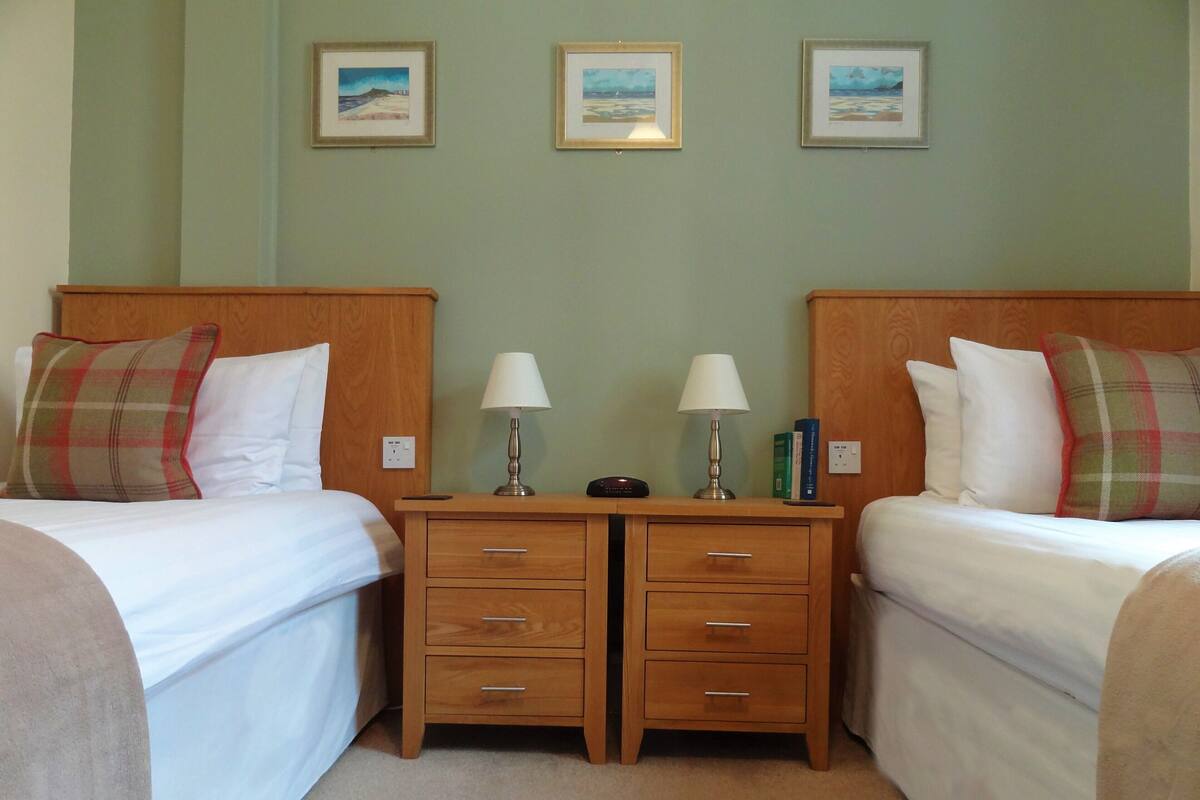 A hotel room with two single beds, a wooden nightstand between them, and a small table with a lamp. There are three framed pictures on the wall above the beds.