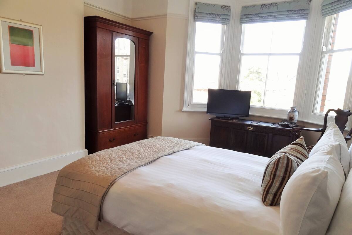 A neatly made bed with a quilt, a wooden dresser with a mirror, a television on a stand, and a window with curtains.
