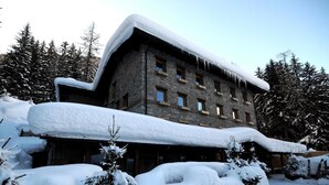 Un edificio de piedra con múltiples ventanas, rodeado de árboles cubiertos de nieve y un techo nevado.