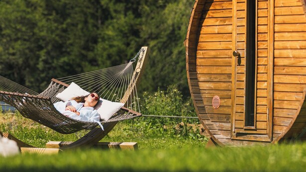 Una cabaña de madera con una puerta circular, una hamaca con una persona acostada y un paisaje montañoso de fondo.