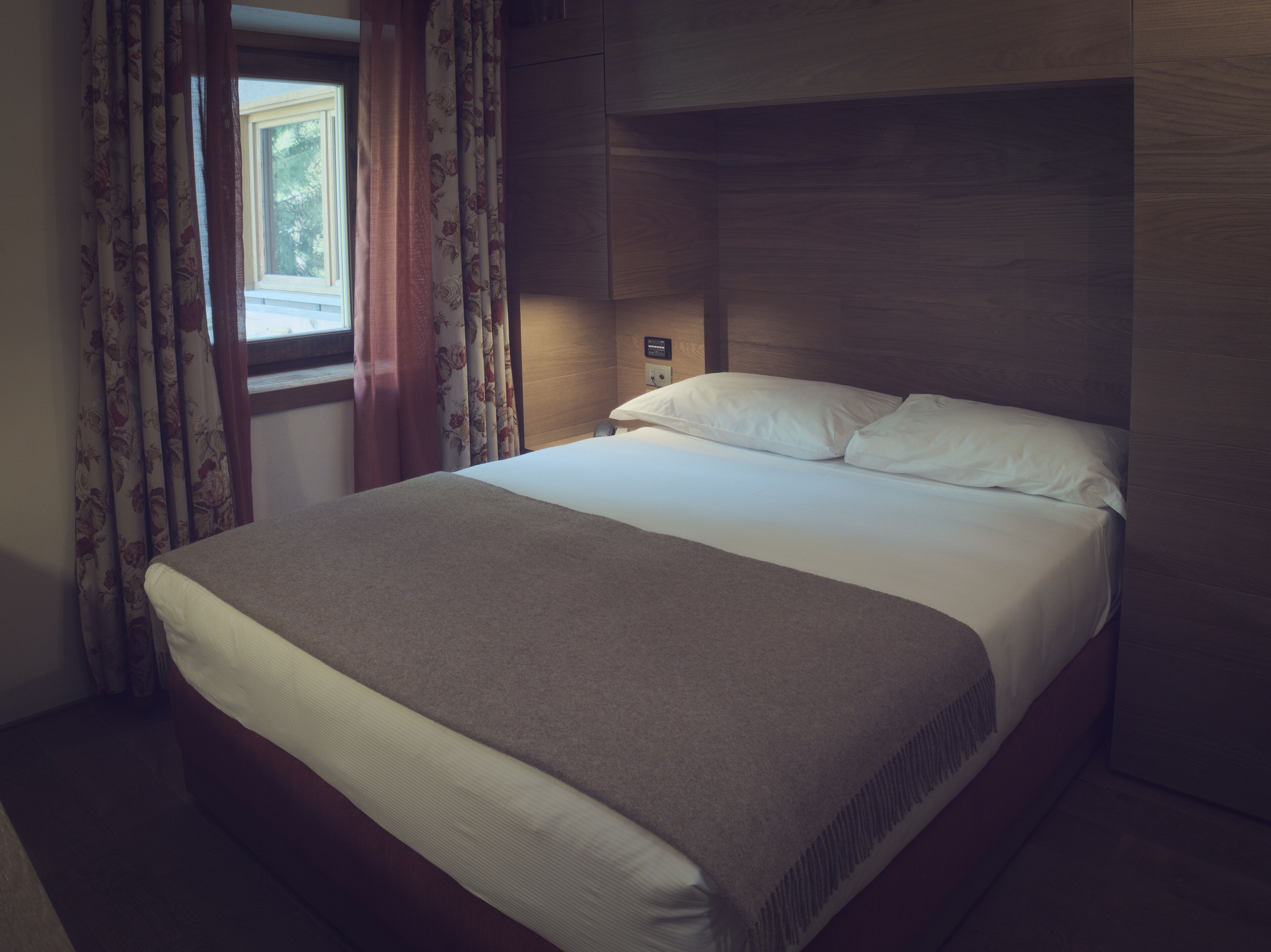A neatly made bed in a hotel room with a wooden headboard, a bedside table, and curtains.