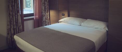 A neatly made bed in a hotel room with a wooden headboard, a bedside table, and curtains.