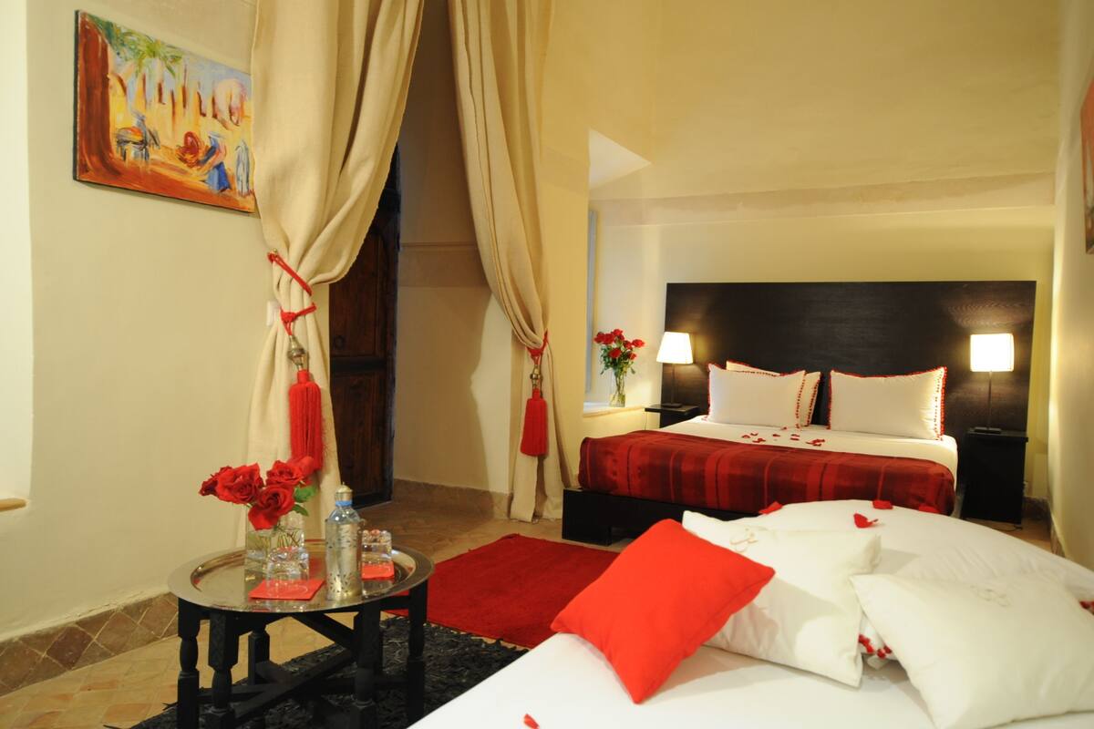 A hotel room with a bed, a bedside table with a vase of flowers, a red throw blanket, and a painting on the wall.
