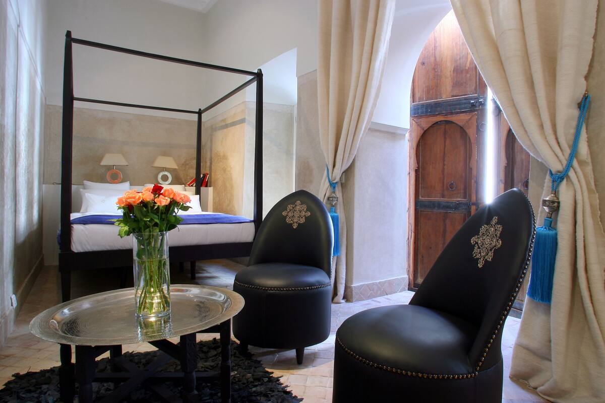 A bedroom with a four-poster bed, a small round table with a vase of flowers, two black leather chairs, and a wooden door with a blue rope.