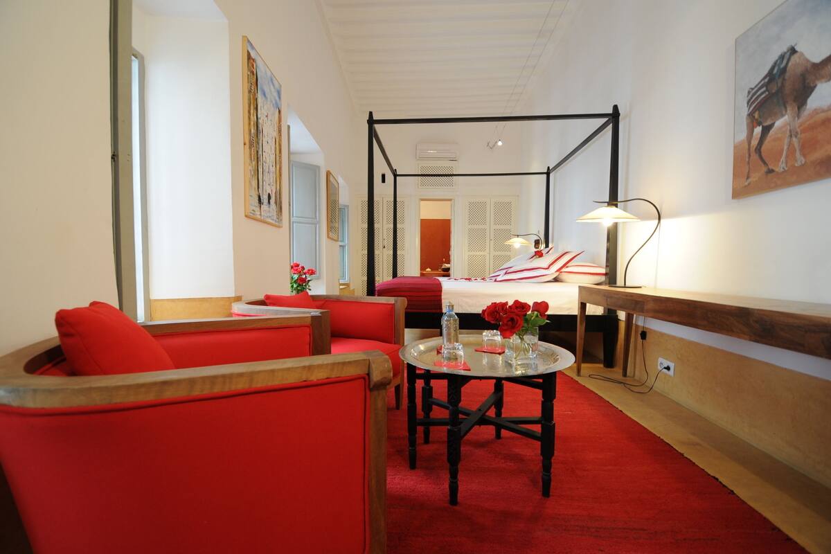 A hotel room with a four-poster bed, red upholstered chairs, a round table with a vase of red roses, and a painting of a camel on the wall.
