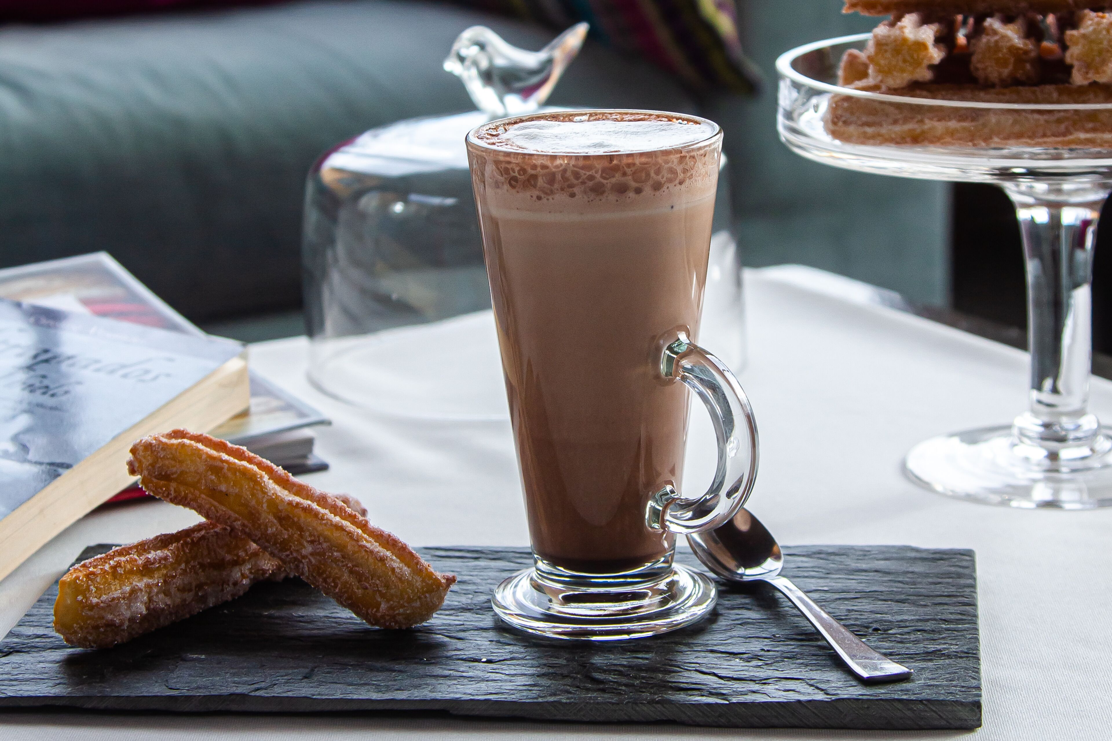 Una taza alta de vidrio con chocolate caliente y asa, dos churros sobre una tabla de pizarra, y una jarra de vidrio al fondo.