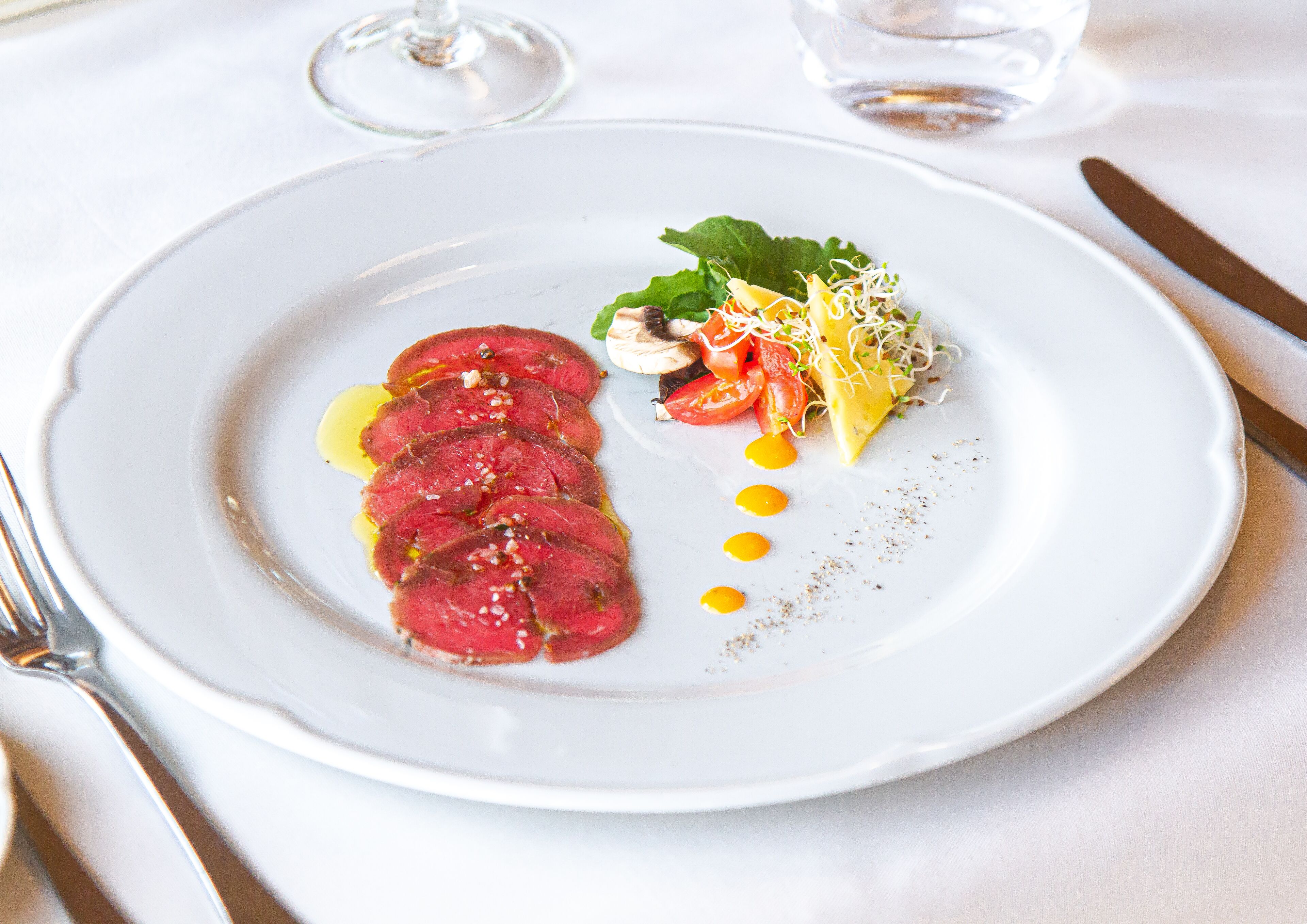 A white plate with a dish of sliced meat, vegetables, and a small salad, accompanied by a glass of water and a wine glass.