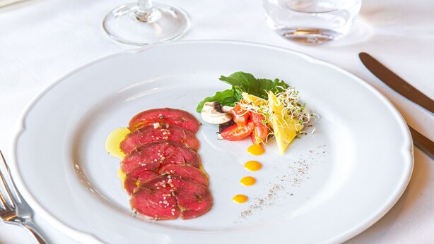 Ein weißer Teller mit einer Portion geschnittenem Fleisch, Gemüse und einem kleinen Salat, dazu ein Glas Wasser und ein Weinglas.