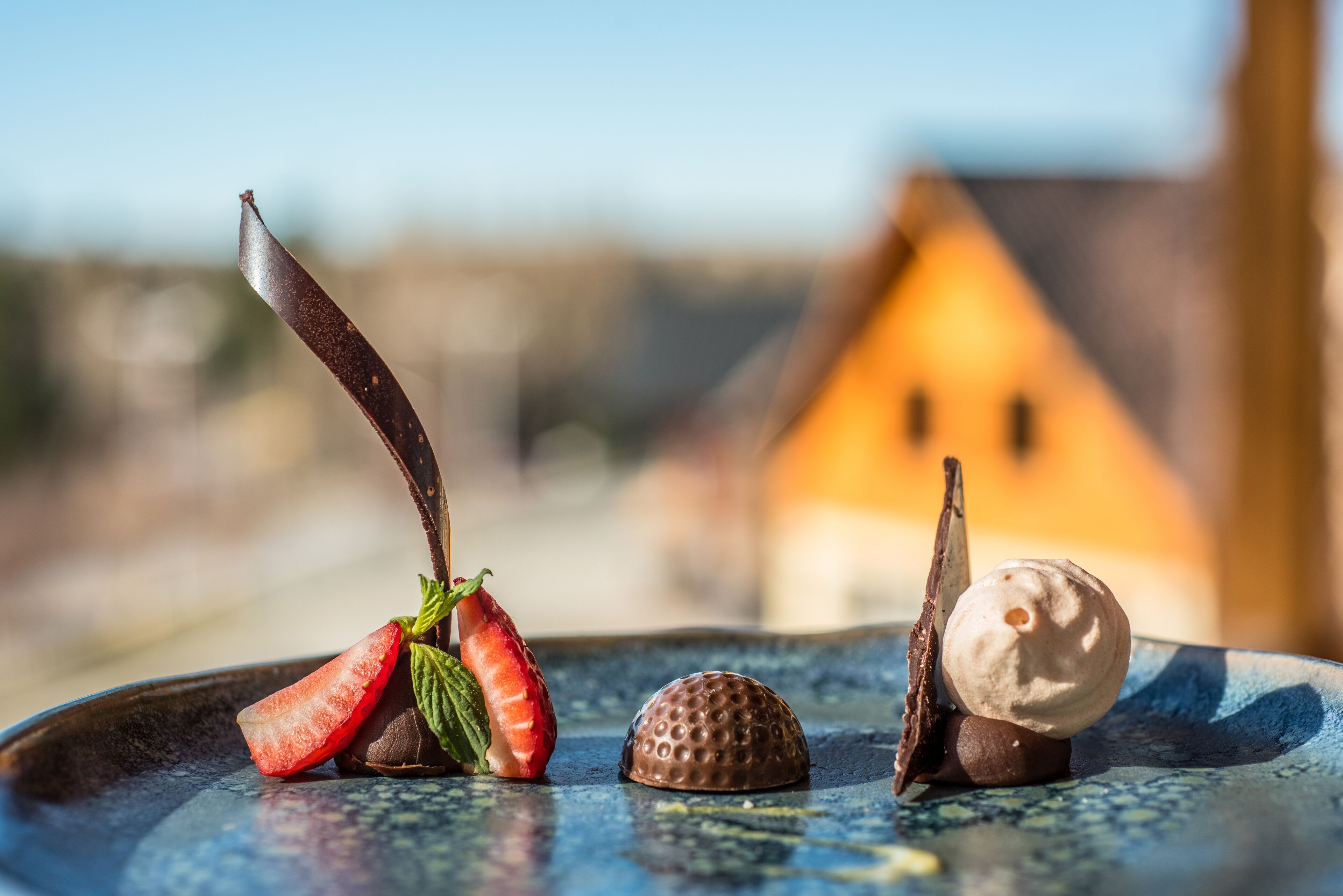 Plat de postre amb xocolata i maduixes sobre una taula, amb un fons borrat d'un edifici.