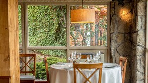 Un coin repas avec une table ronde dressée pour quatre personnes, entourée de chaises en bois, et offrant une vue sur la verdure grâce à de grandes fenêtres.