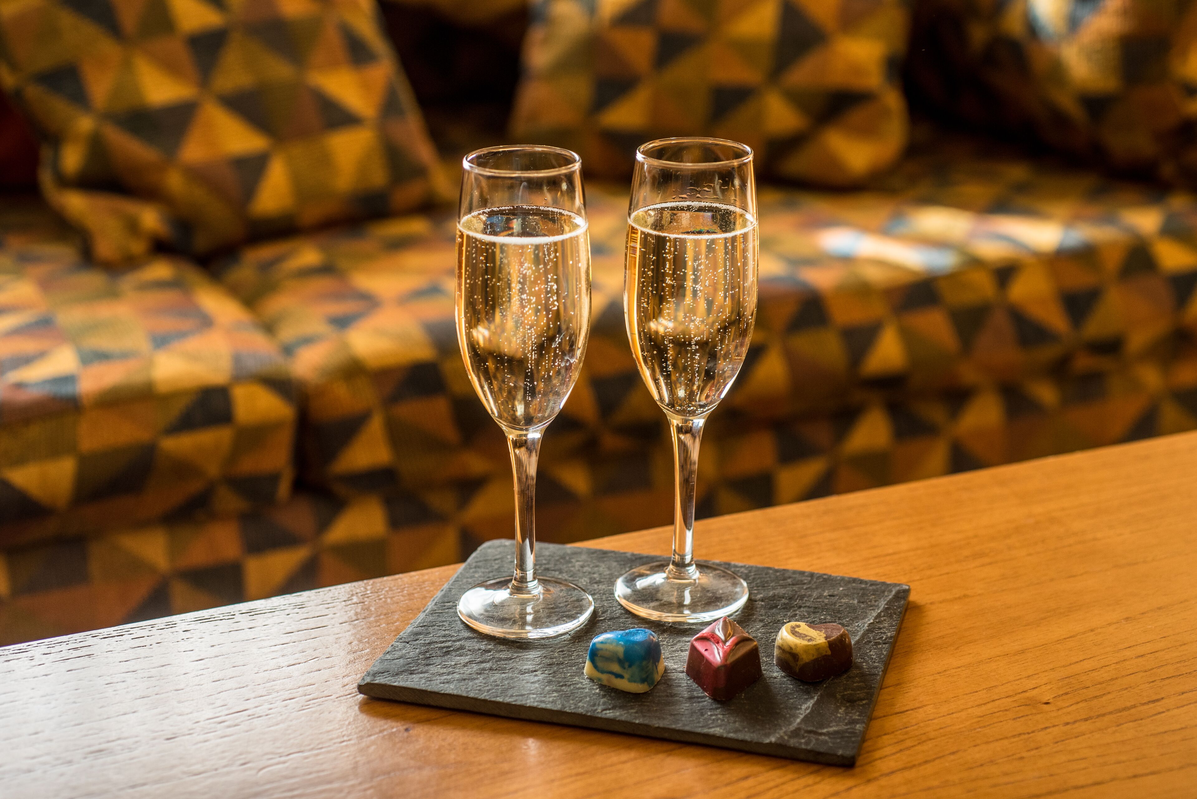 Two glasses of champagne on a slate coaster, with a small plate of chocolates in front.