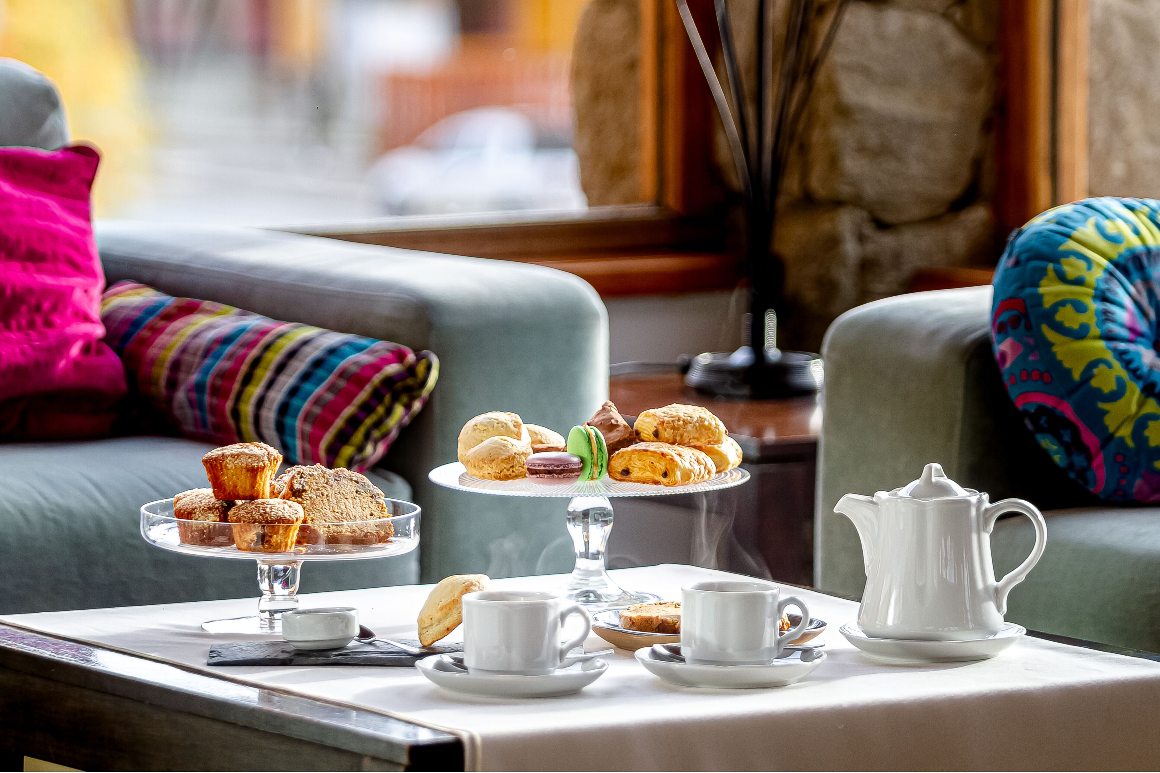 A table with pastries, tea cups, and a teapot, set on a windowsill with a view of a stone wall and colorful cushions.