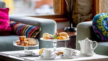 A table with pastries, tea cups, and a teapot, set on a windowsill with a view of a stone wall and colorful cushions.