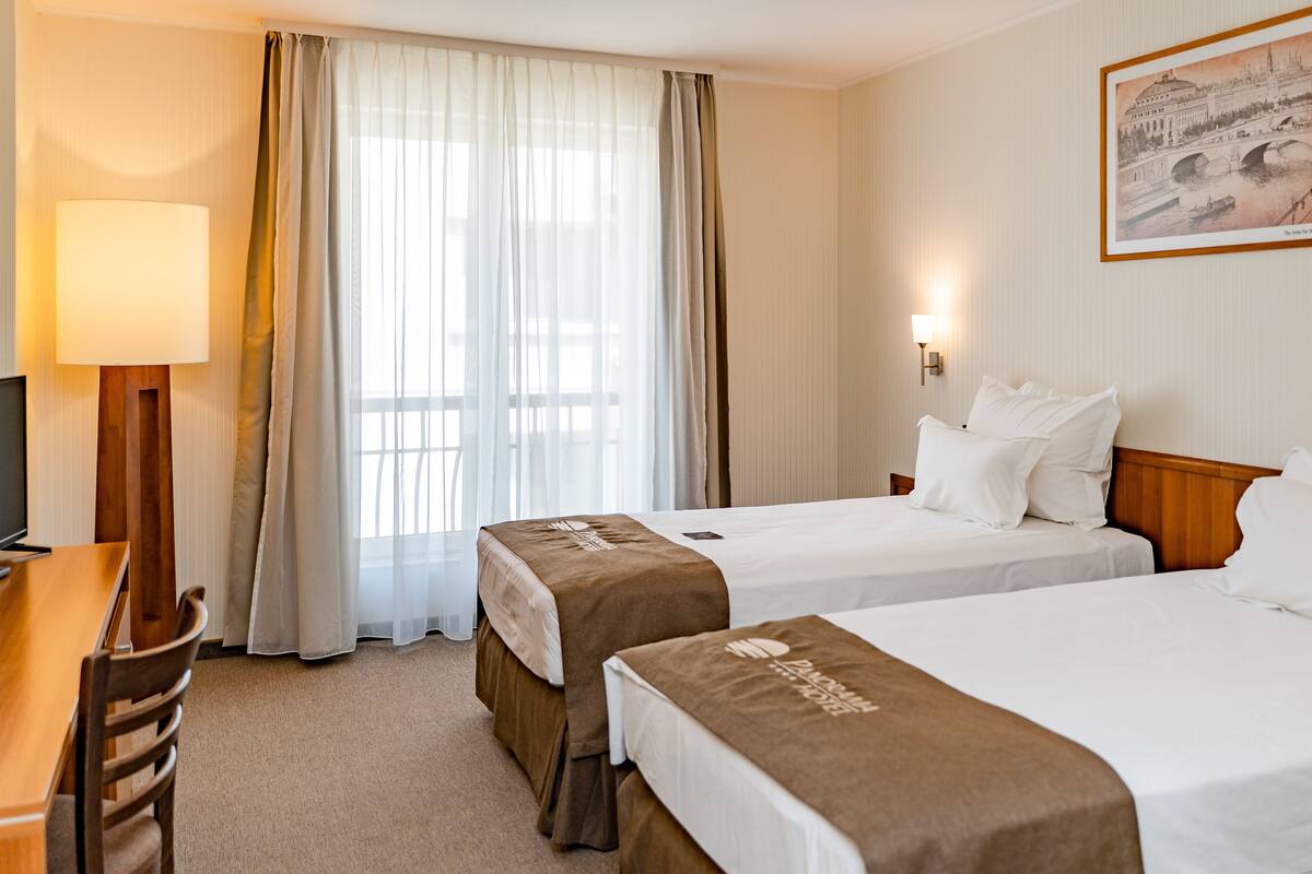 A hotel room with two beds, a desk, a chair, a lamp, and a window with curtains.
