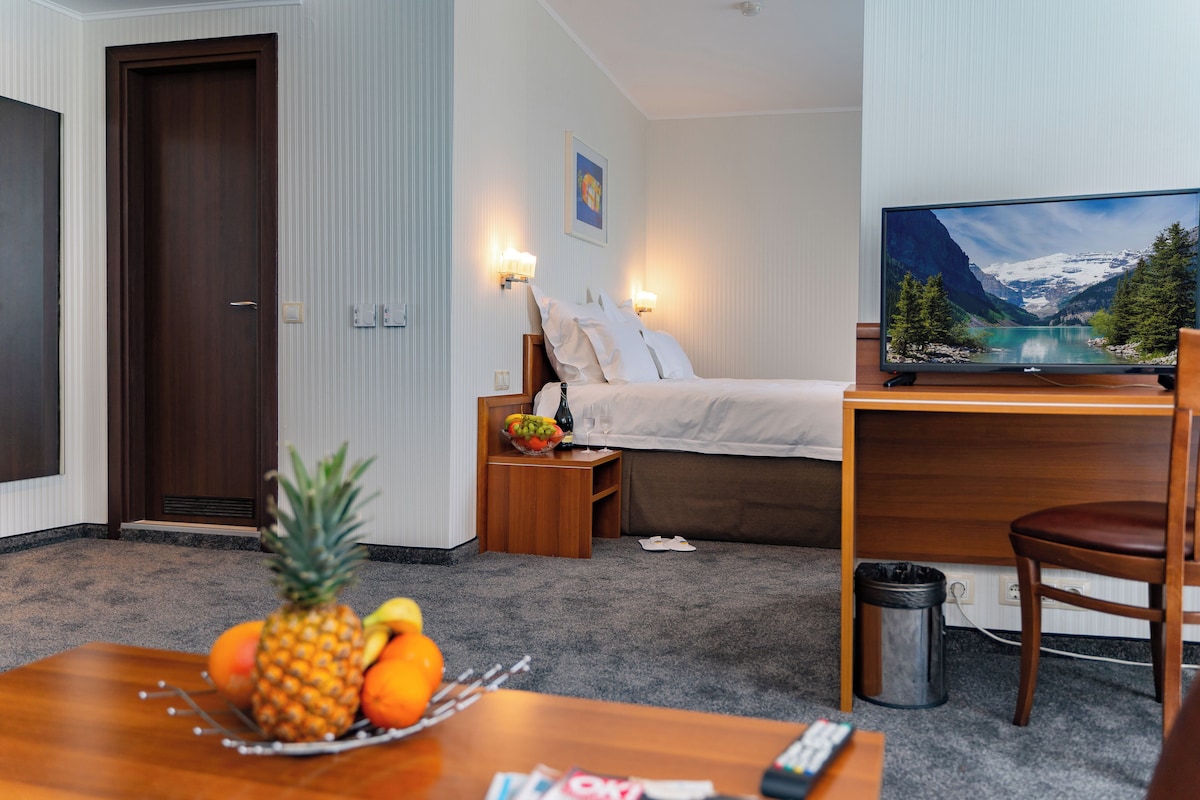 Habitación de hotel con cama, lámparas de noche, televisor, escritorio, silla y frutero sobre la mesa.