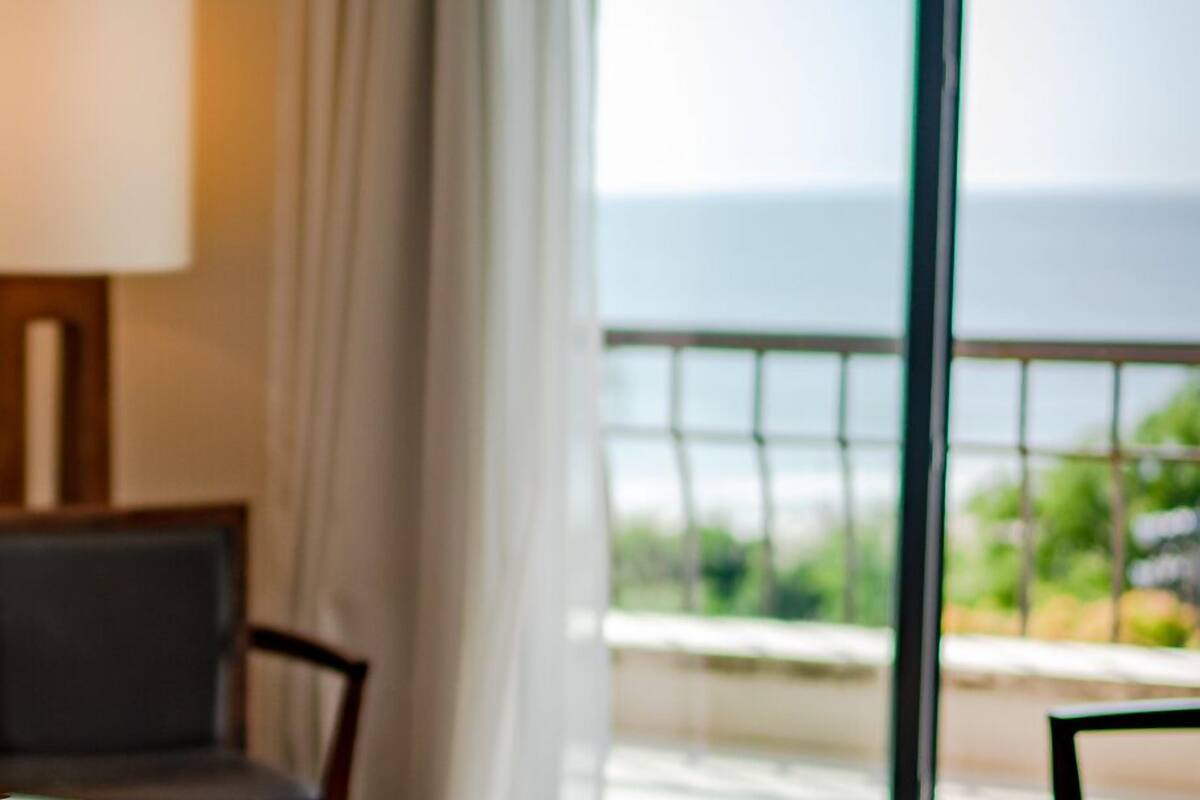 A room with a view of the sea, a window with curtains, and a table with a lamp.