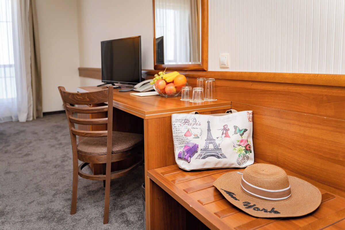 A hotel room with a wooden desk, a chair, a computer, a glass of water, a fruit bowl, a decorative bag, and a straw hat.