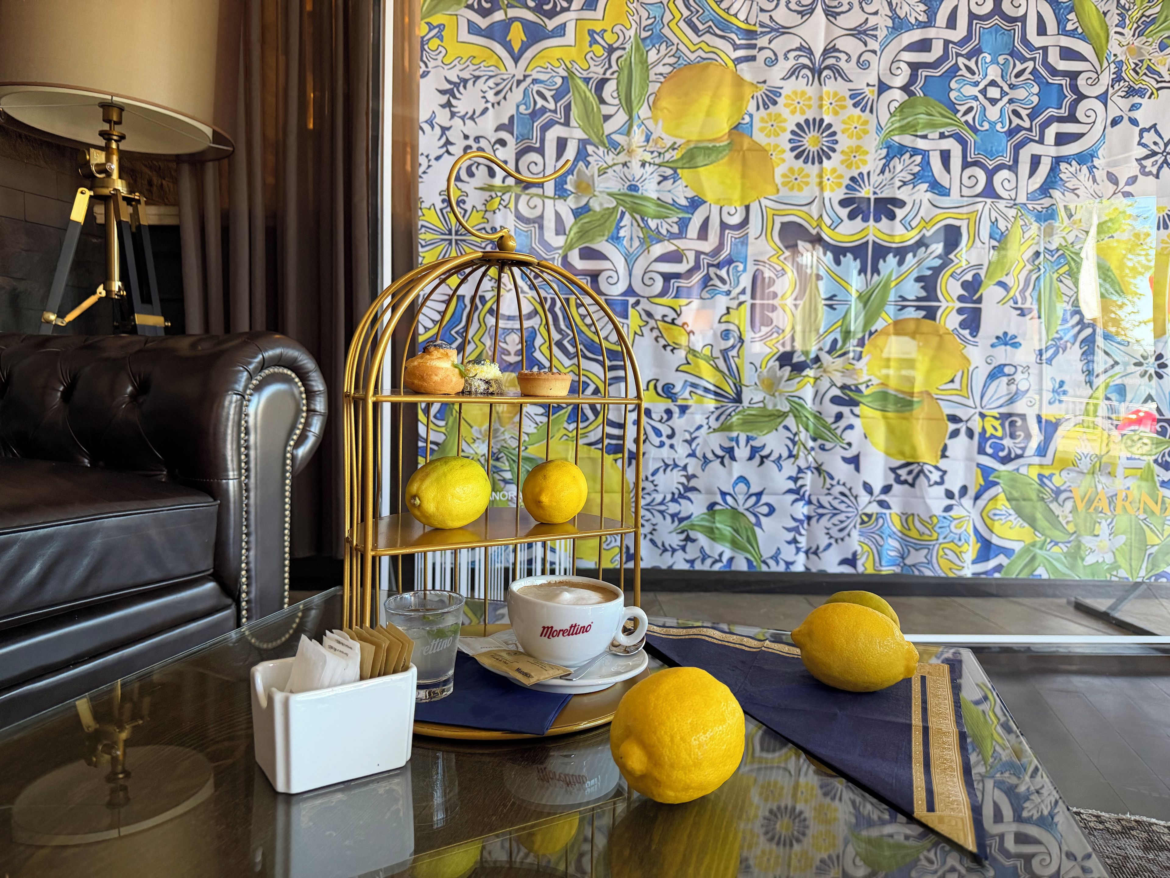 A room with a black leather sofa, a glass coffee table with a white cup and saucer, a gold birdcage with lemons, and a patterned wall.