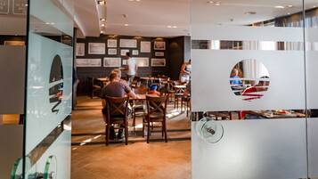 A modern restaurant interior with wooden tables and chairs, framed pictures on the walls, and a glass partition with a circular cutout.