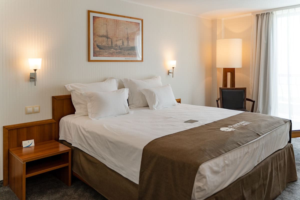 A hotel room with a large bed, two bedside tables, a chair, a painting on the wall, and two wall-mounted lamps.