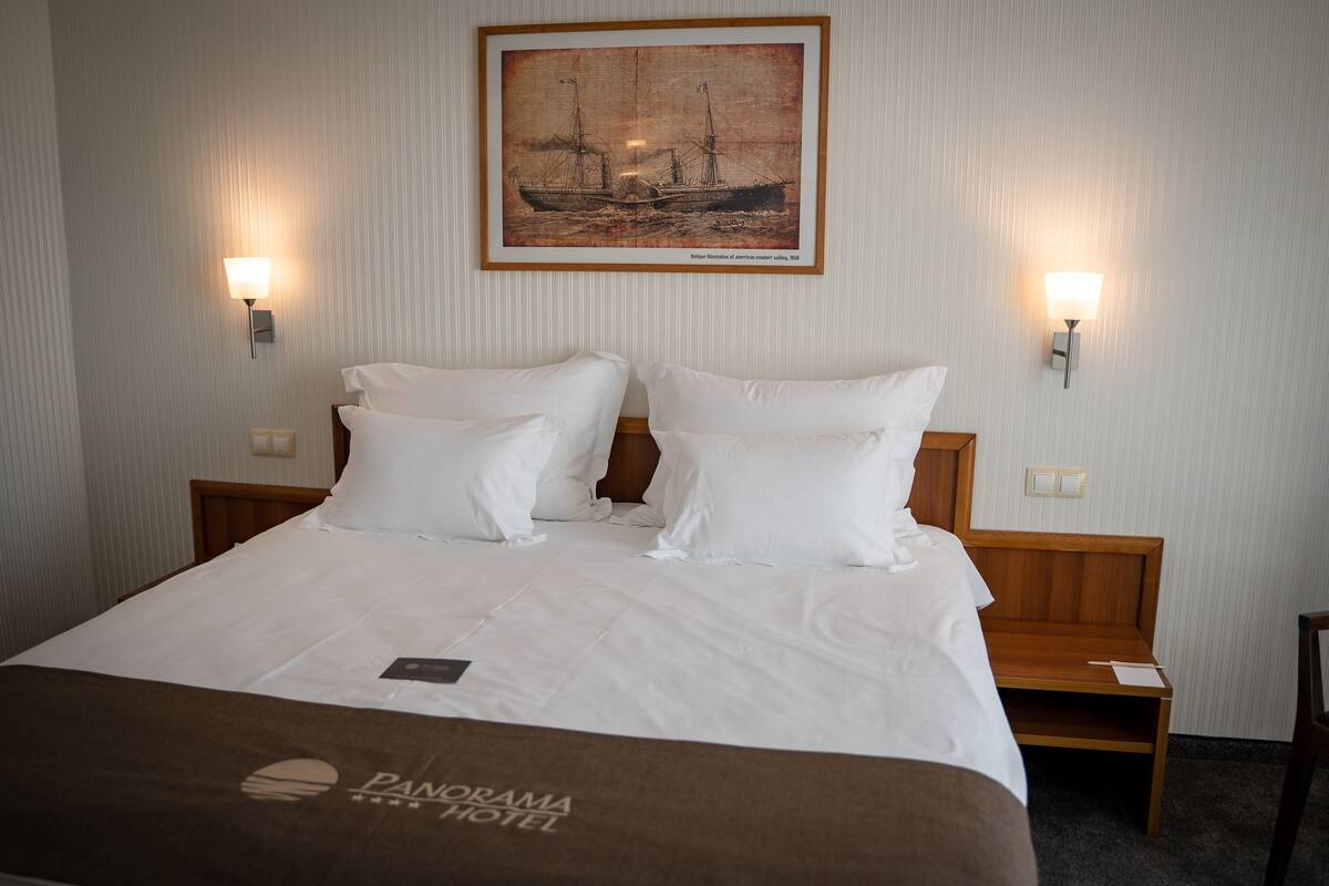 A hotel room with a bed, two bedside lamps, a framed picture of a sailing ship, and a brown blanket with the hotel logo.