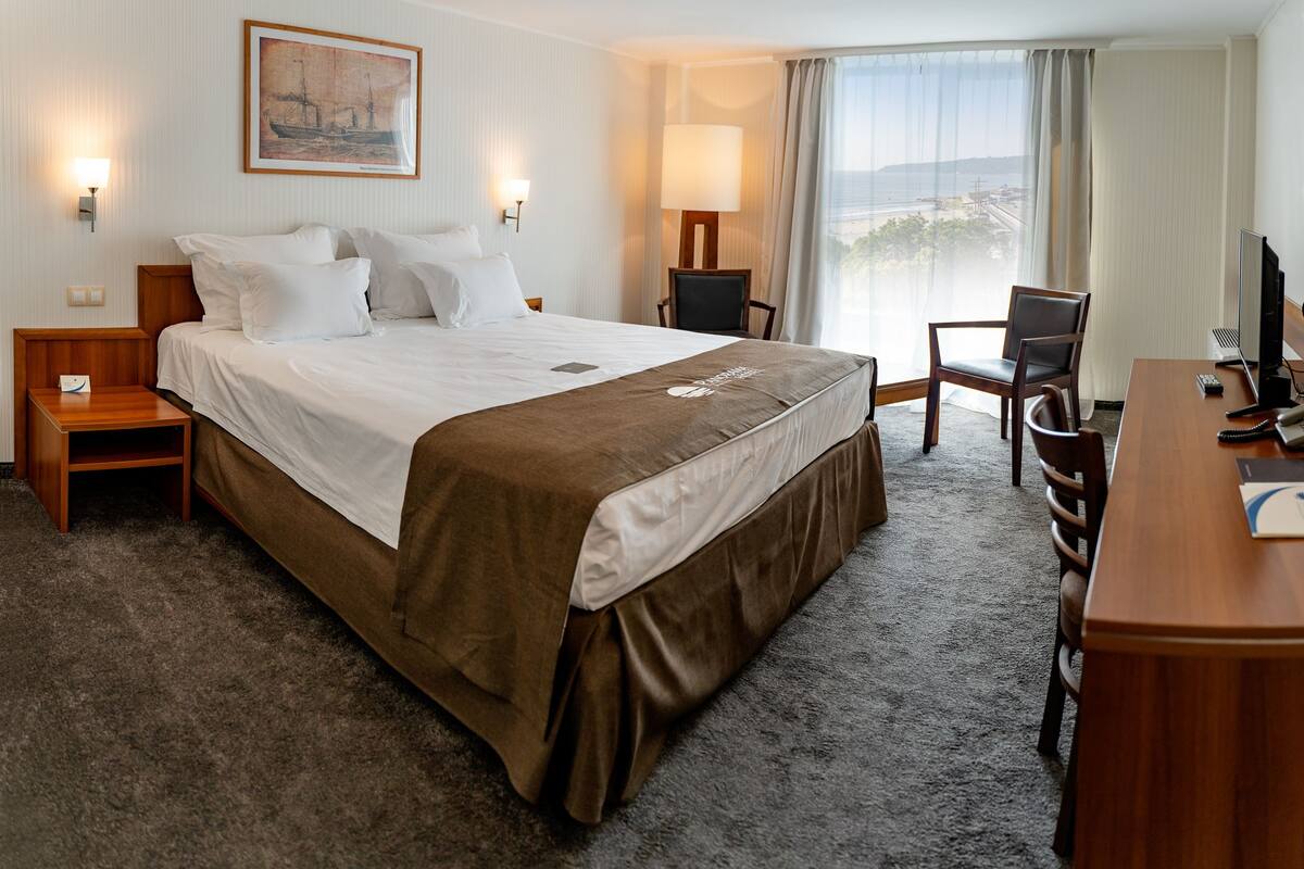A hotel room with a large bed, a desk with a computer, a chair, and a view of the outdoors.