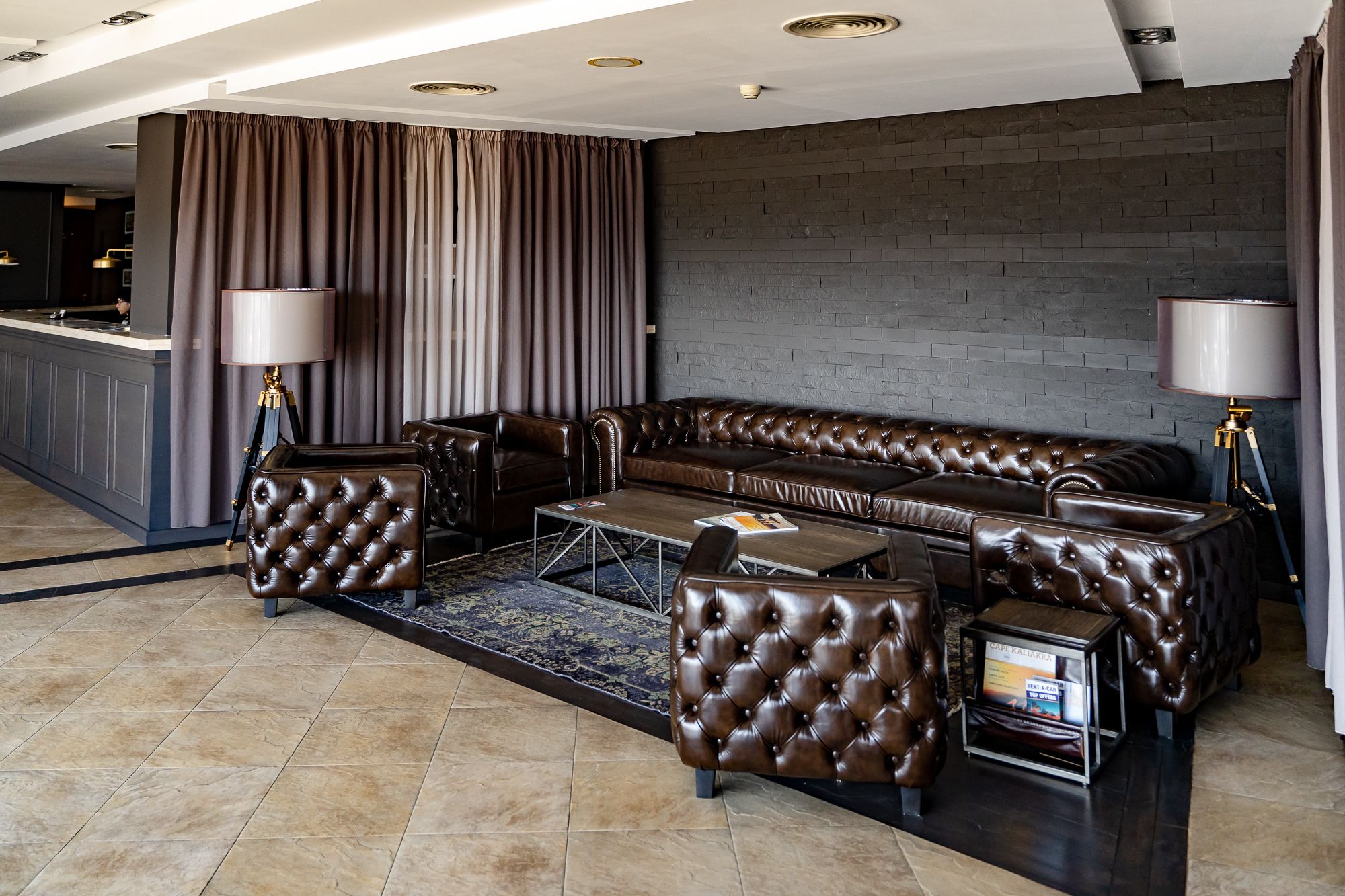 A luxurious living room with a dark brown leather sofa, matching armchairs, and a coffee table. There are two floor lamps and a bar area in the background.