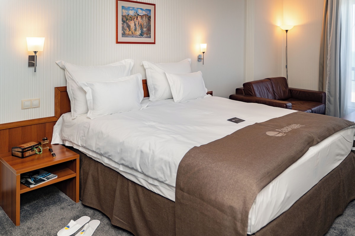 Una habitación de hotel con una cama grande, una mesita de noche de madera, un sillón de cuero marrón y dos lámparas fijadas a la pared.