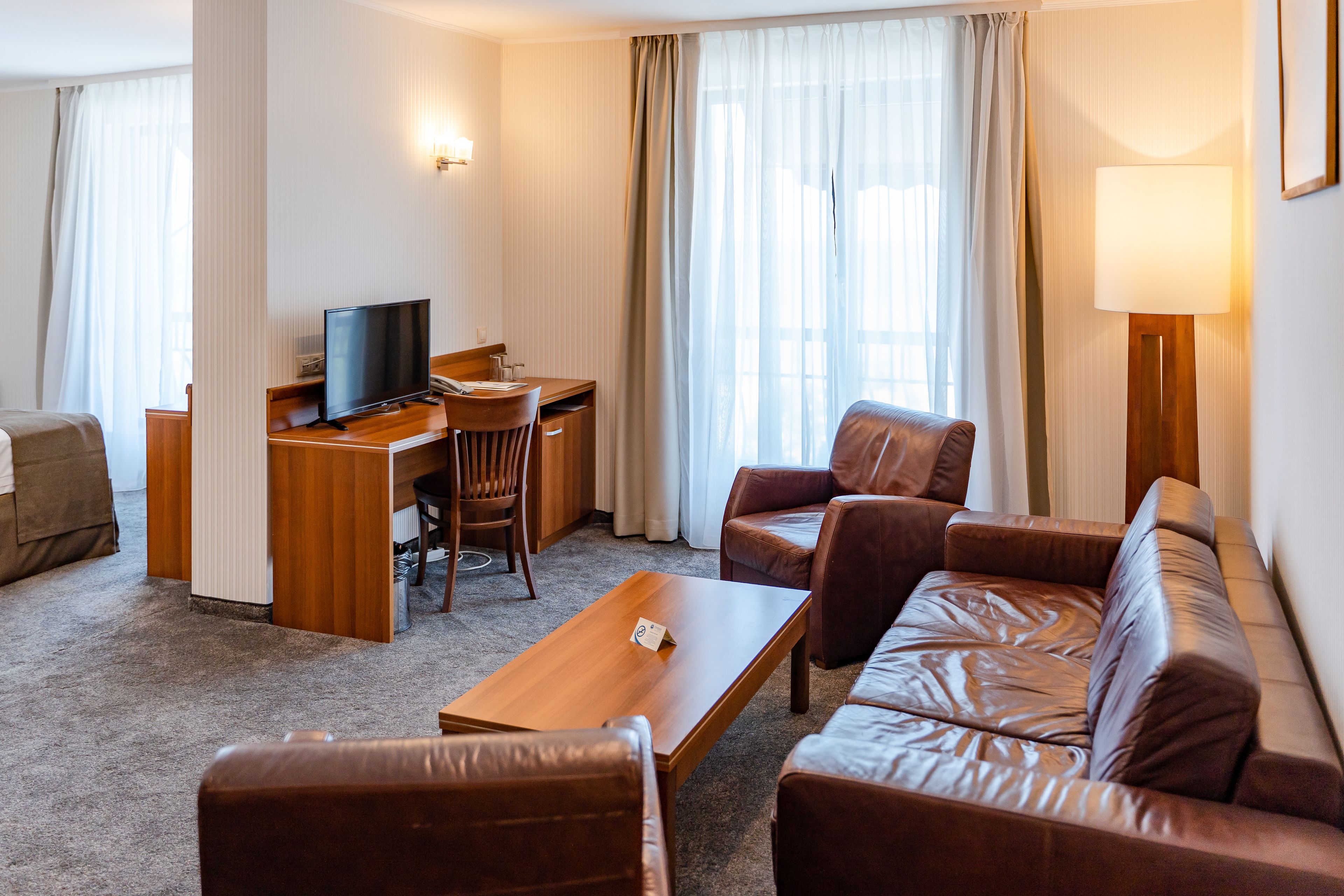 Habitación de hotel con sofá, escritorio, televisor, lámpara y ventana con cortinas.