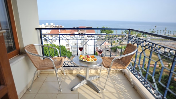 A balcony with a table set for two, two wicker chairs, and a view of the sea and buildings.