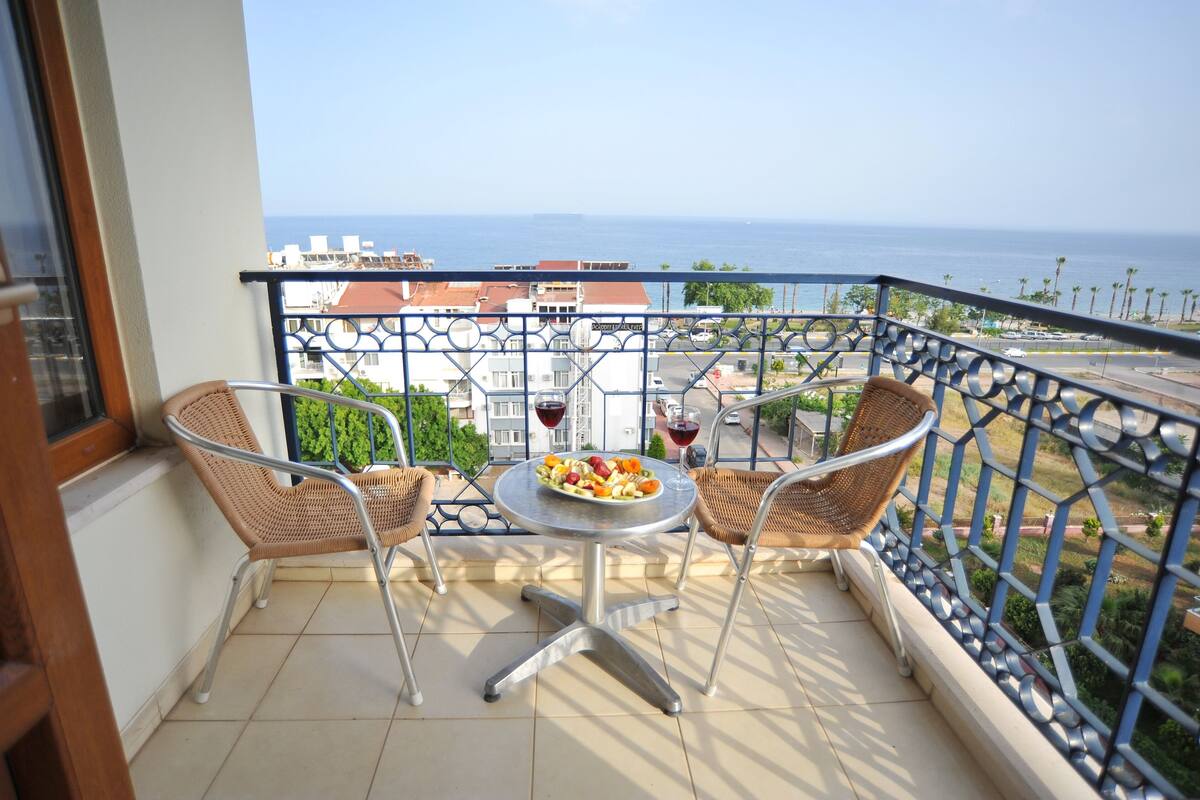 Un balcón con una mesa para dos, dos sillas de mimbre y vista al mar y edificios.