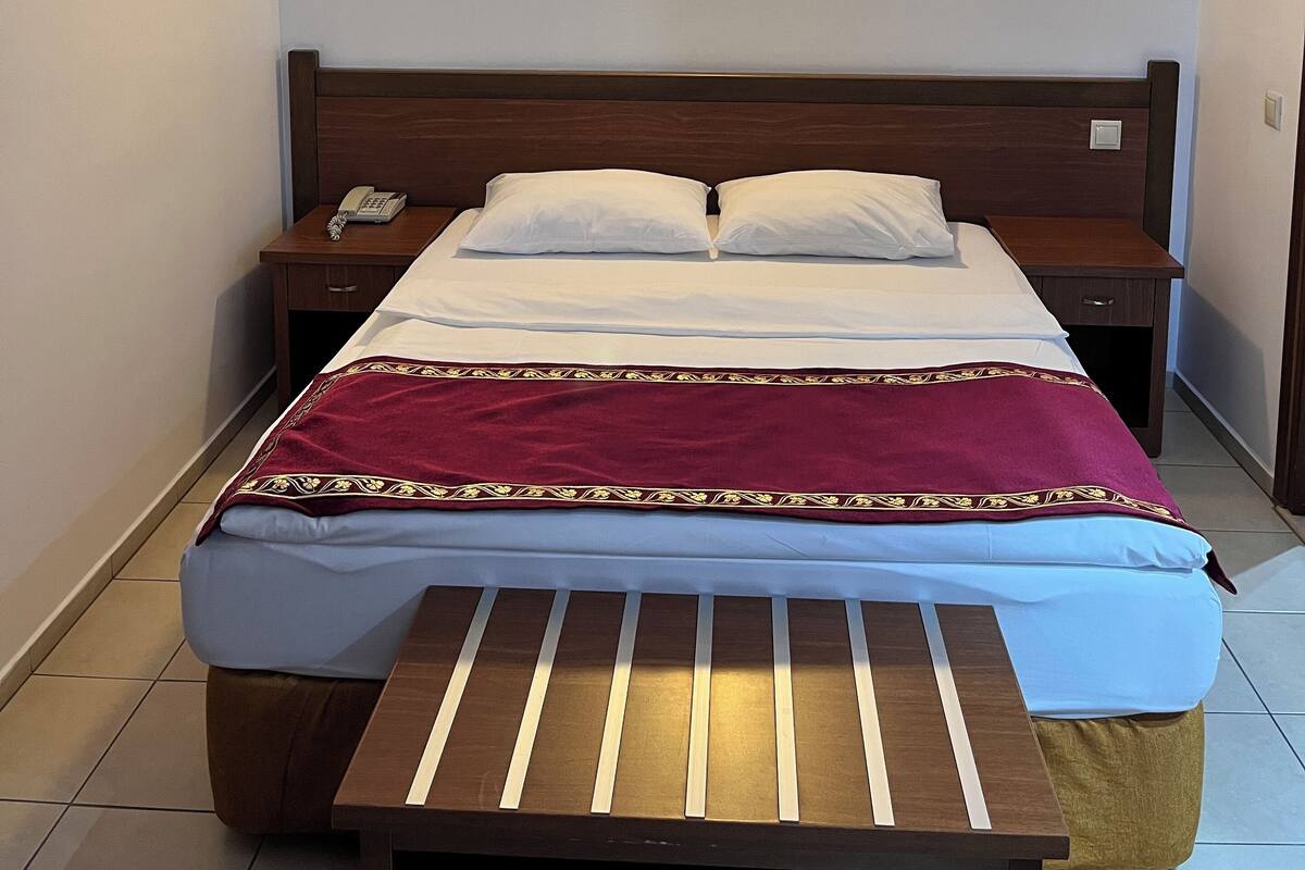 Una cama bien hecha con cabecera de madera, una mesita de noche con teléfono y una lámpara fijada a la pared.