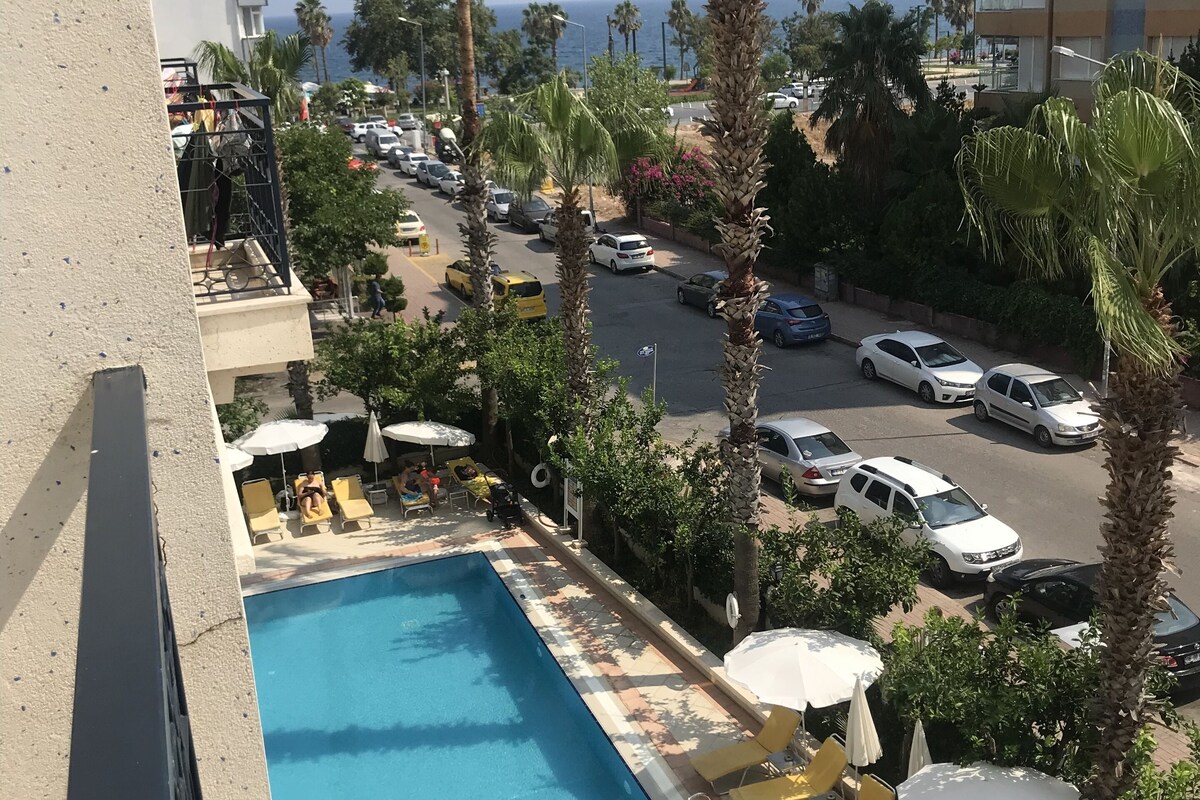 A swimming pool with lounge chairs and umbrellas, surrounded by palm trees and a view of the street and ocean.