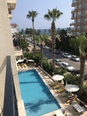Une piscine entourée de palmiers, avec des chaises longues et des parasols, d’où l’on peut admirer la rue et l’océan.