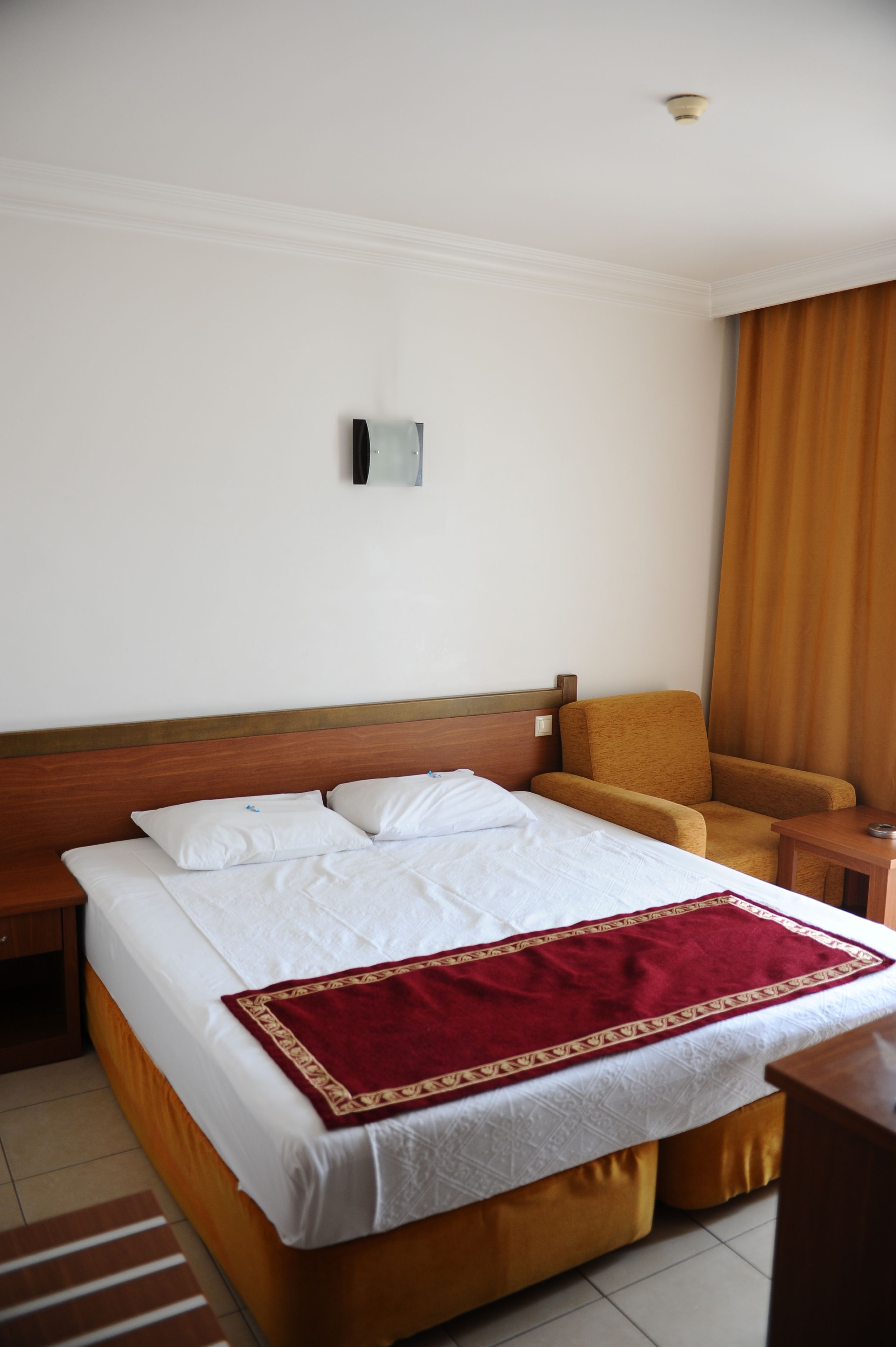 A hotel room with a large bed, a wooden headboard, a red throw blanket, a wooden nightstand, and a chair.