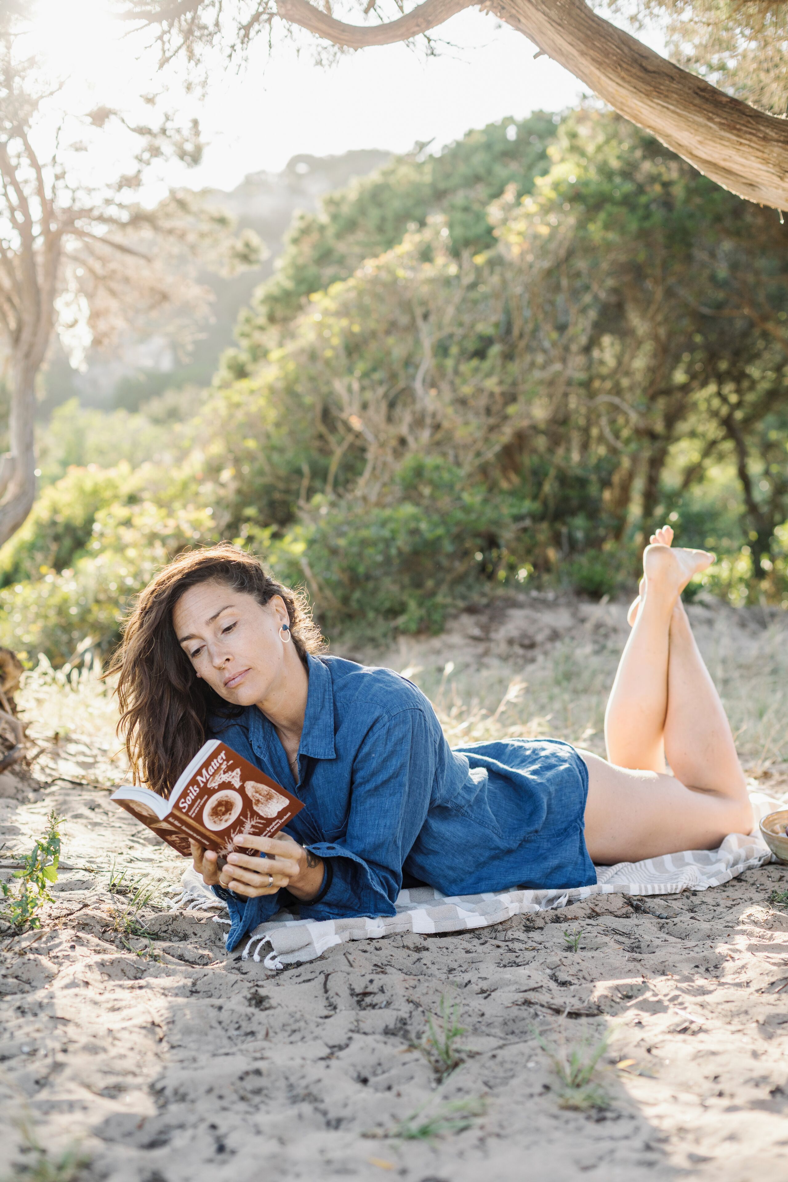 Eine Person liegt auf einer Decke unter einem Baum und liest ein Buch.