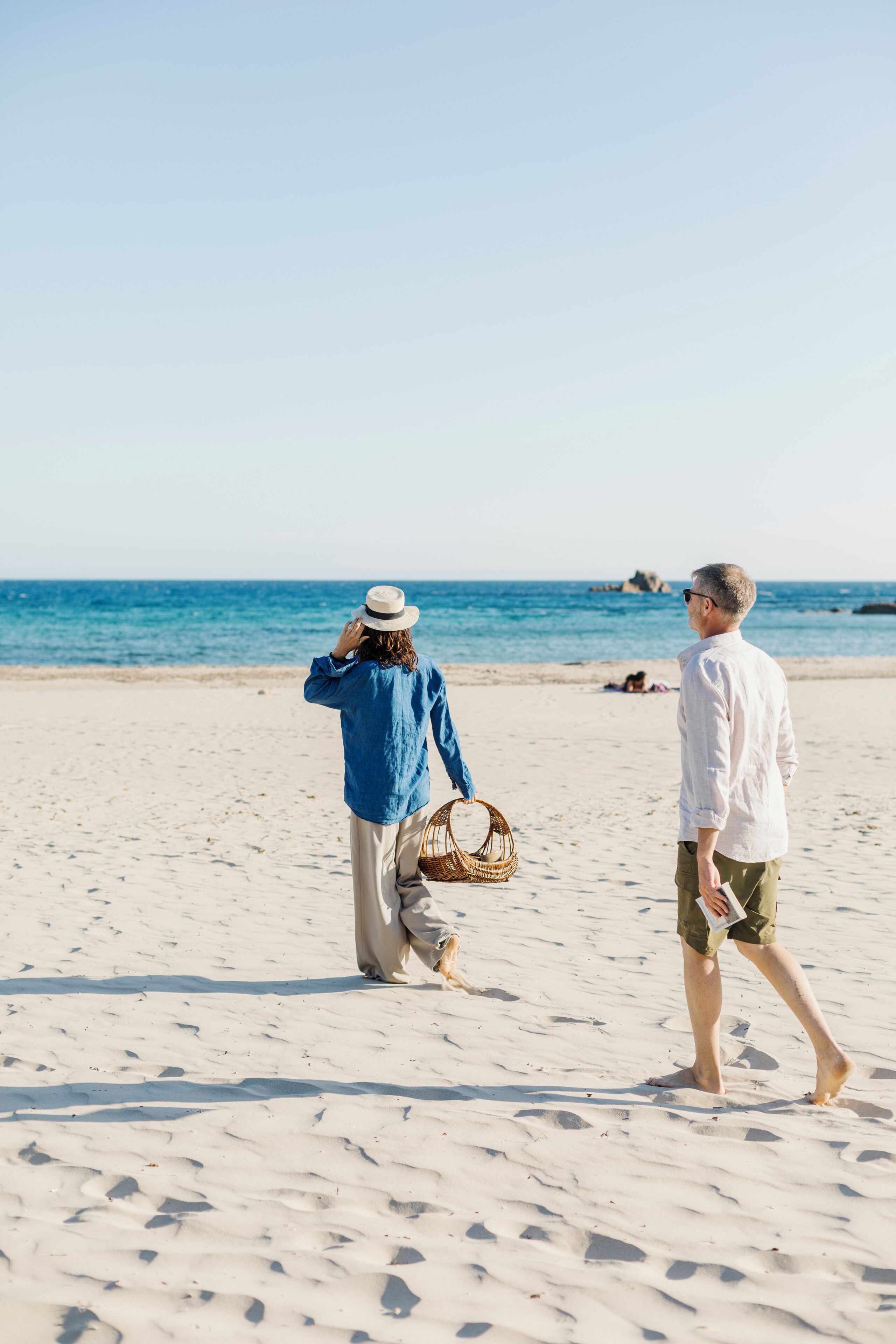 Une plage de sable où deux personnes se promènent, l’une d’elles portant un panier.