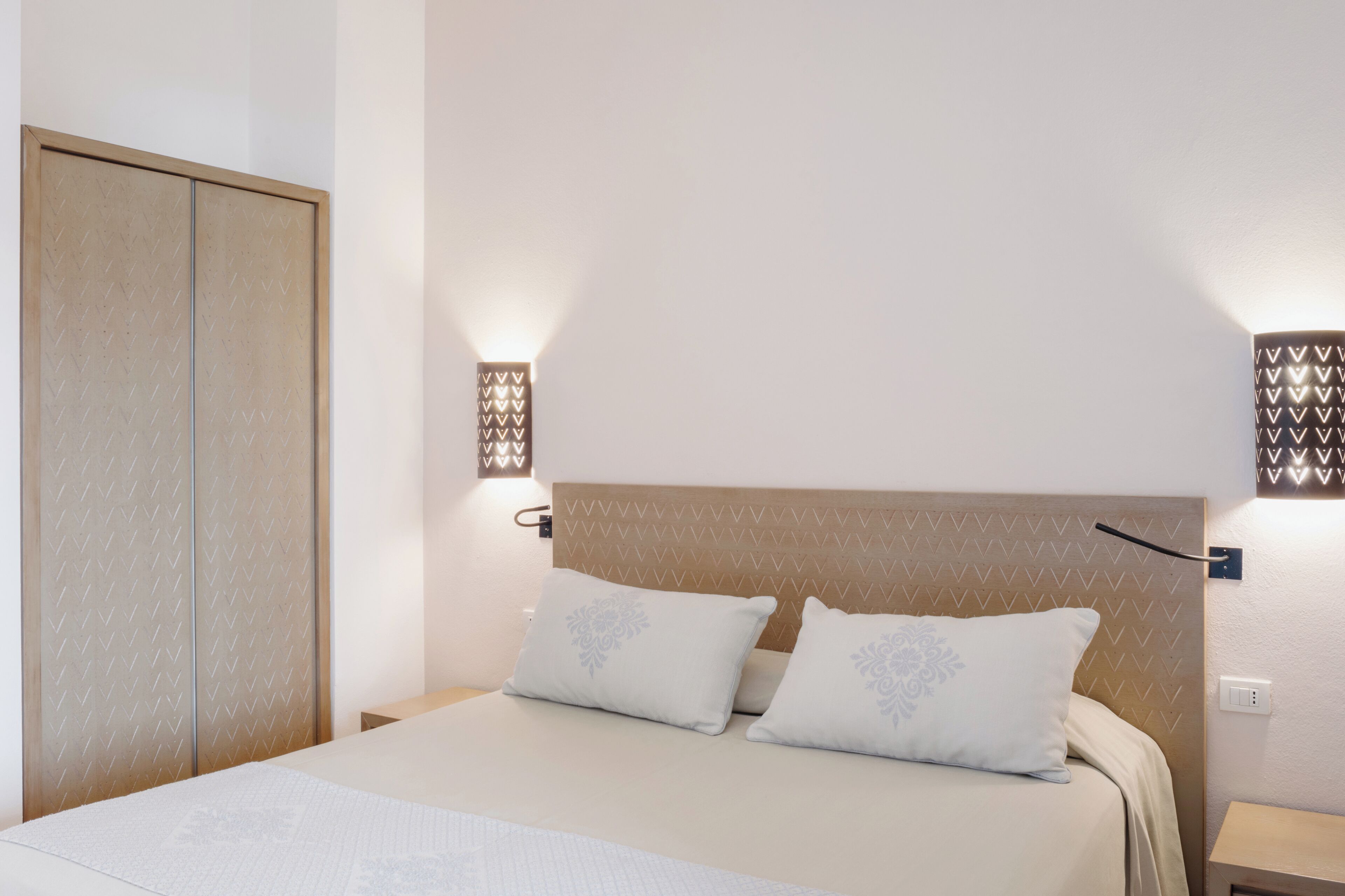 Een opgemaakt bed met hoofdbord, twee bedlampen en een kledingkast.