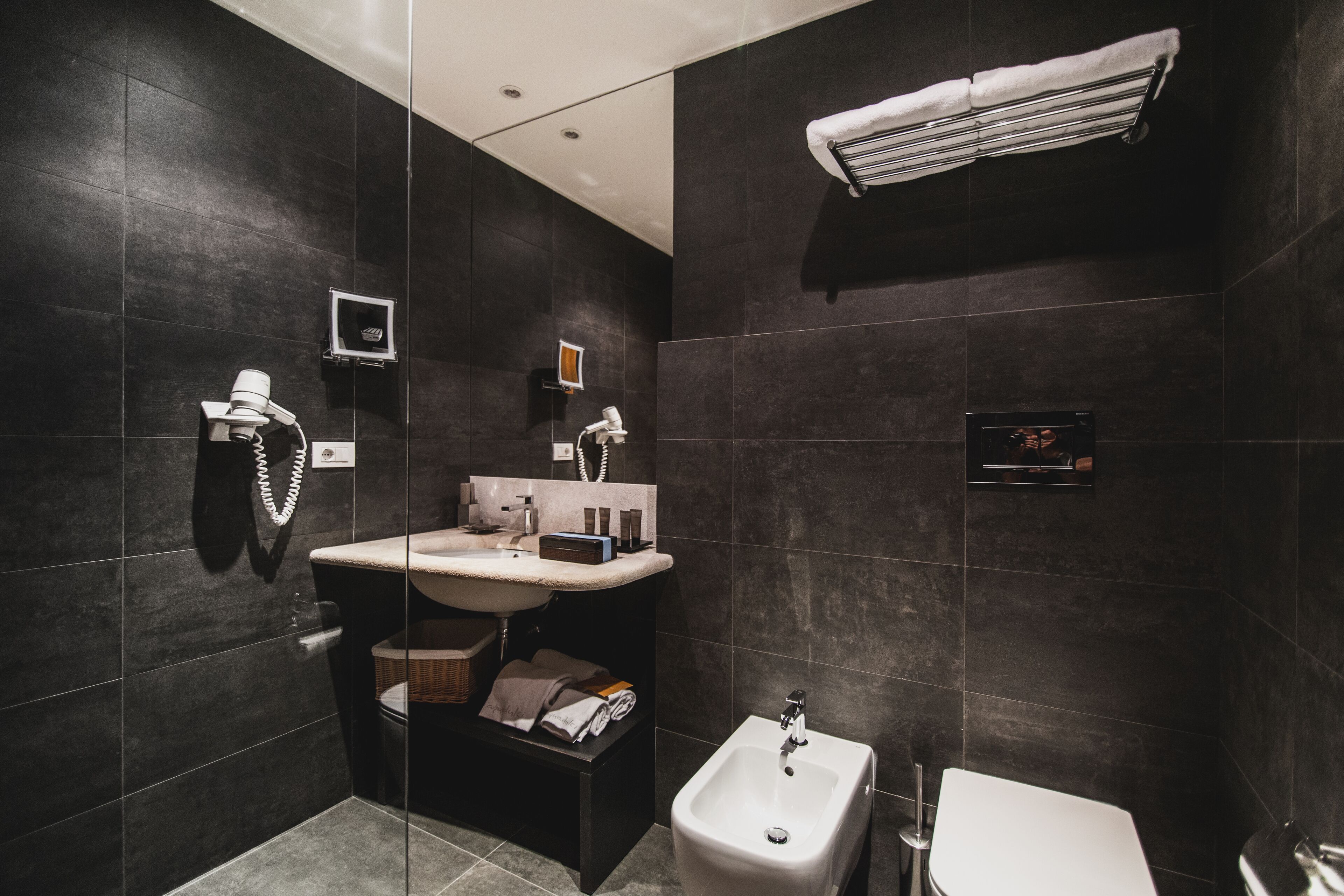 A modern bathroom with dark tiles, a white sink, a bidet, and a towel rack.