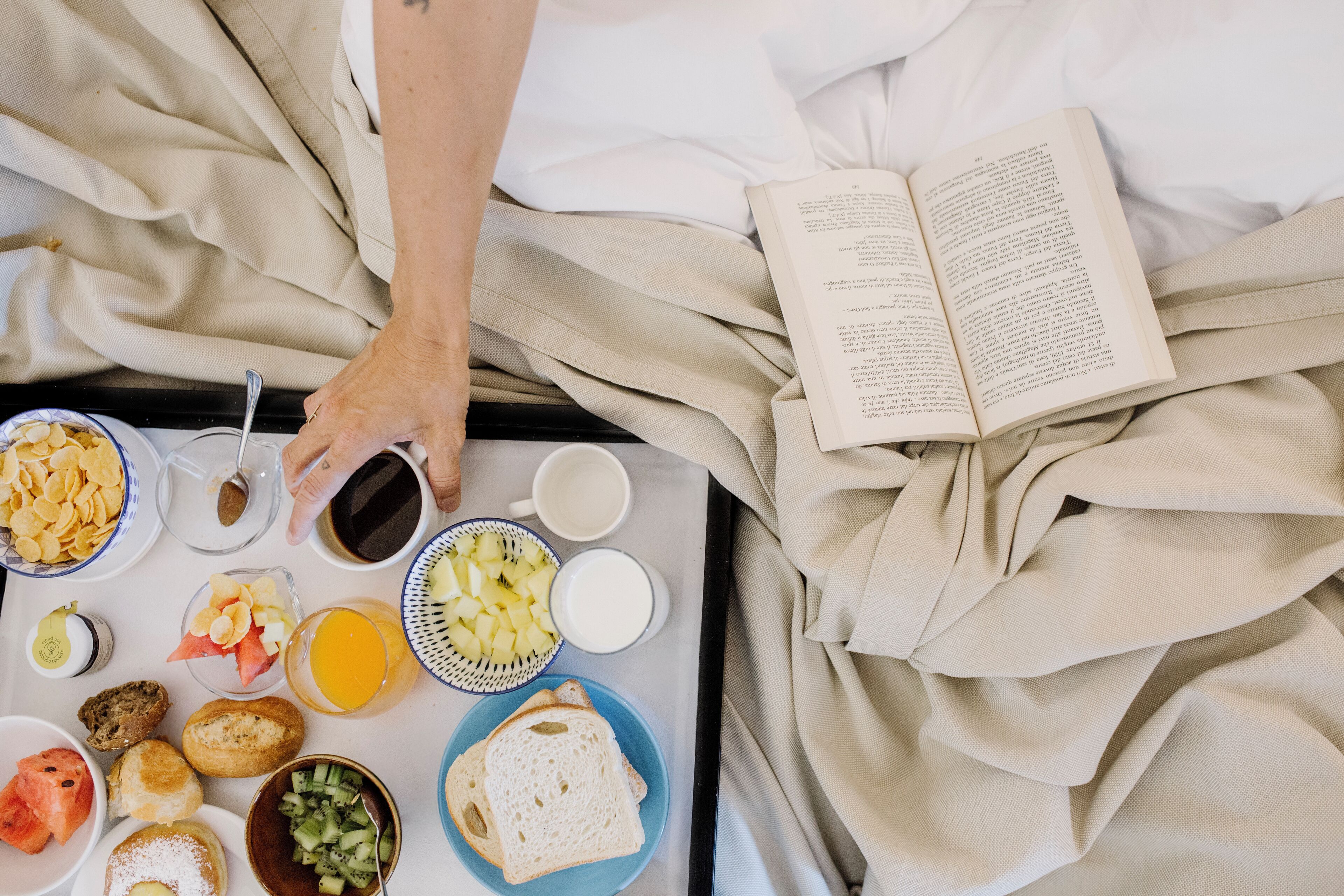 A breakfast tray with various food items and a book on a bed.