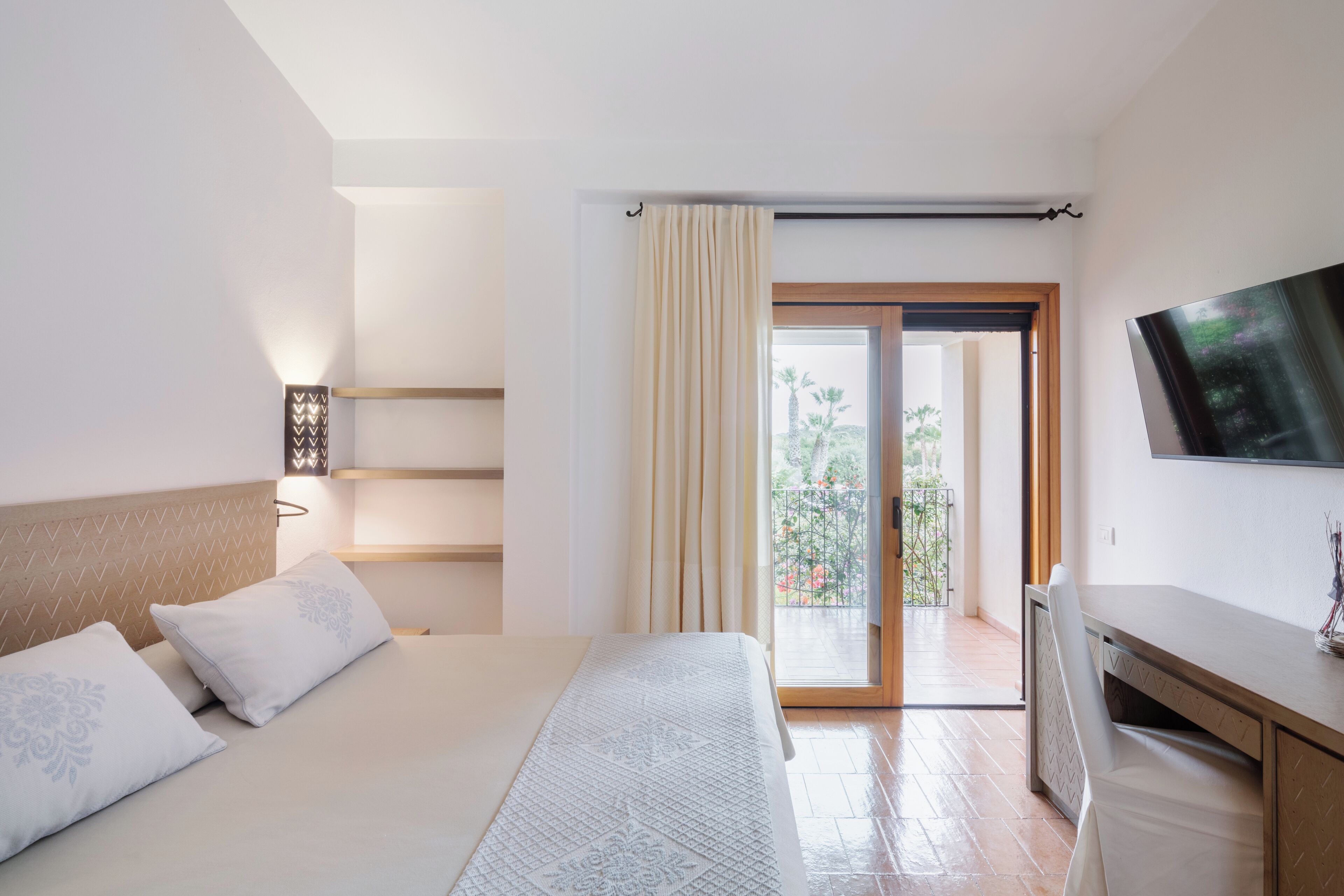 A bedroom with a bed, bedside tables, a desk, a TV, and a balcony with curtains.