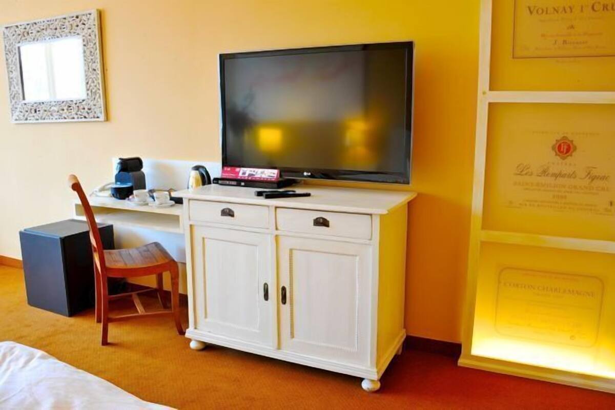 Habitación de hotel con una televisión sobre un mueble blanco, una silla, una mesita con cafetera y diplomas enmarcados en la pared.