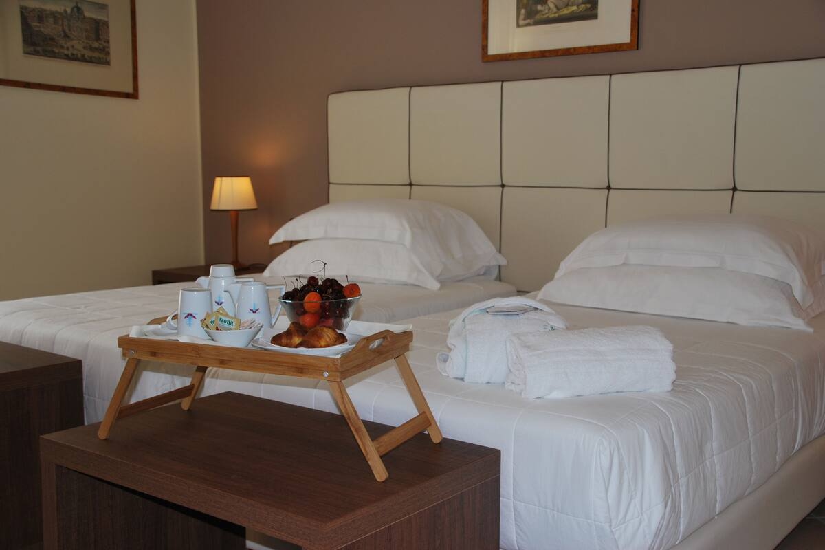 A hotel room with two beds, a wooden side table with a tray of fruit and beverages, and a lamp on the nightstand.